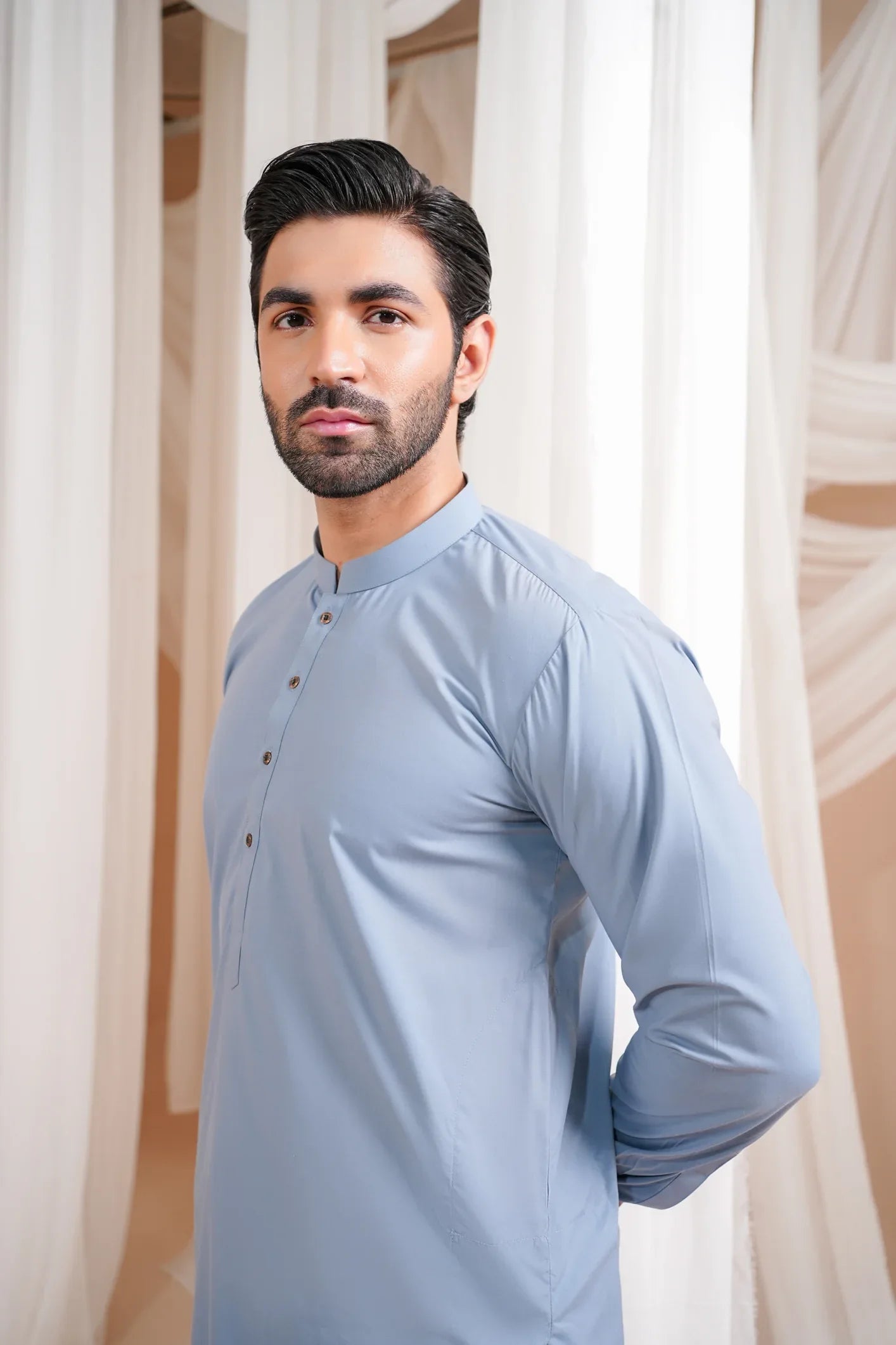 A man wearing a blue luxury blended kurta shalwar, standing against a curtain backdrop.