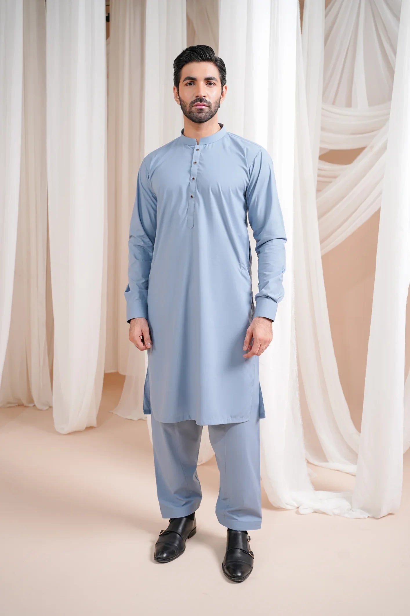 A man wearing a blue luxury blended kurta shalwar, standing against a curtain backdrop.