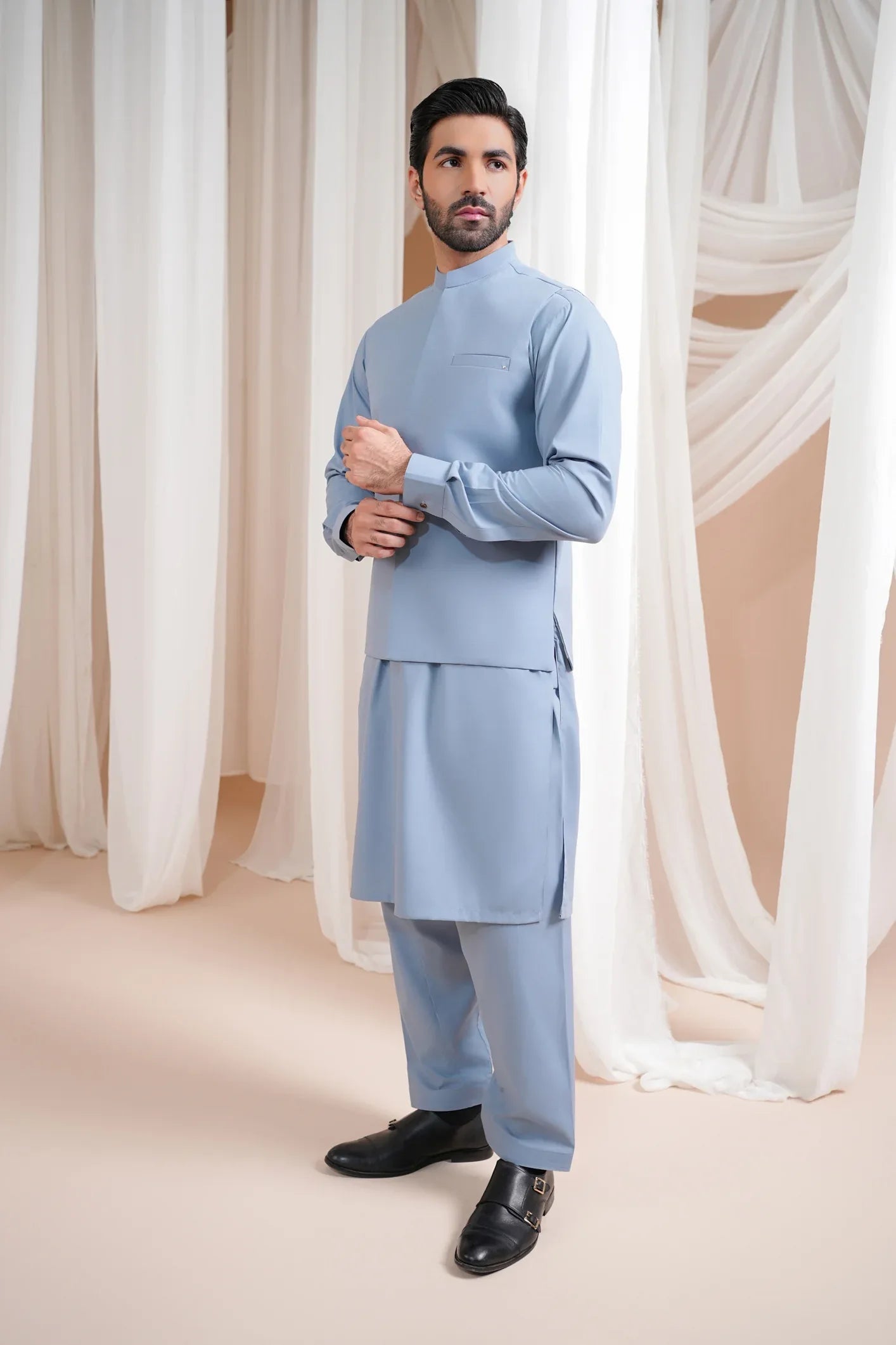 A man wearing a sapphire blue luxury formal kameez shalwar with a blended waistcoat, standing in a room with white drapes and a mandarin collar.