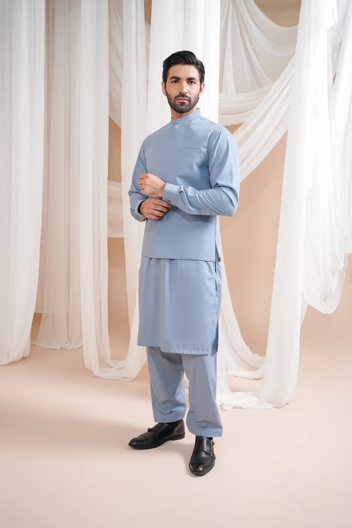 A man wearing a burgundy luxury formal kameez shalwar with a blended waistcoat and full sleeves. He is standing in a room with light curtains behind him.