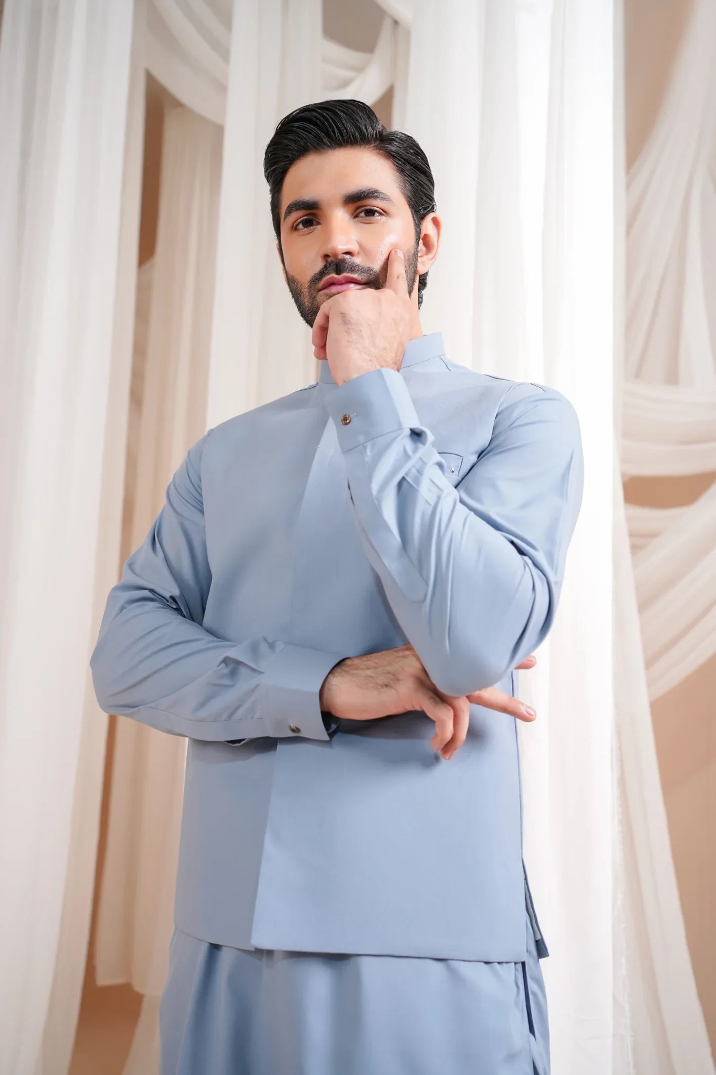 A man wearing a burgundy luxury formal kameez shalwar with a blended waistcoat and full sleeves. He is standing in a room with light curtains behind him.