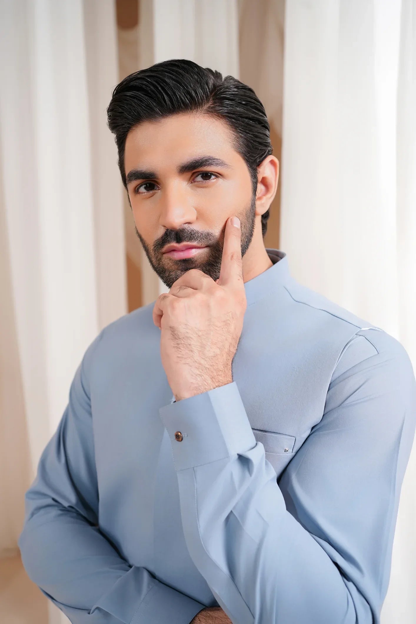 A man wearing a straight cut waistcoat in a sapphire blue color with a mandarin collar and full sleeves, standing in a room with white drapes.