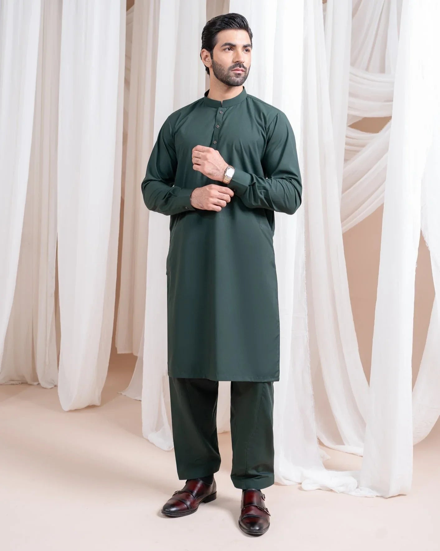 A man standing in a shalwar kameez with a mandarin collar, full sleeves, and a straight kurta and trouser. The background features white drapes and a soft grey background.