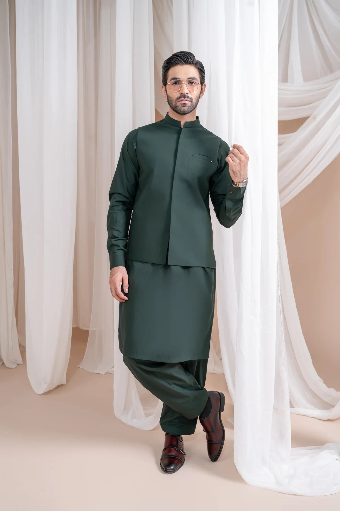 A man wearing a bottle green luxury formal Kameez Shalwar with a patch pocket waistcoat and full sleeves. The outfit is straight cut and suited for all-season wear.