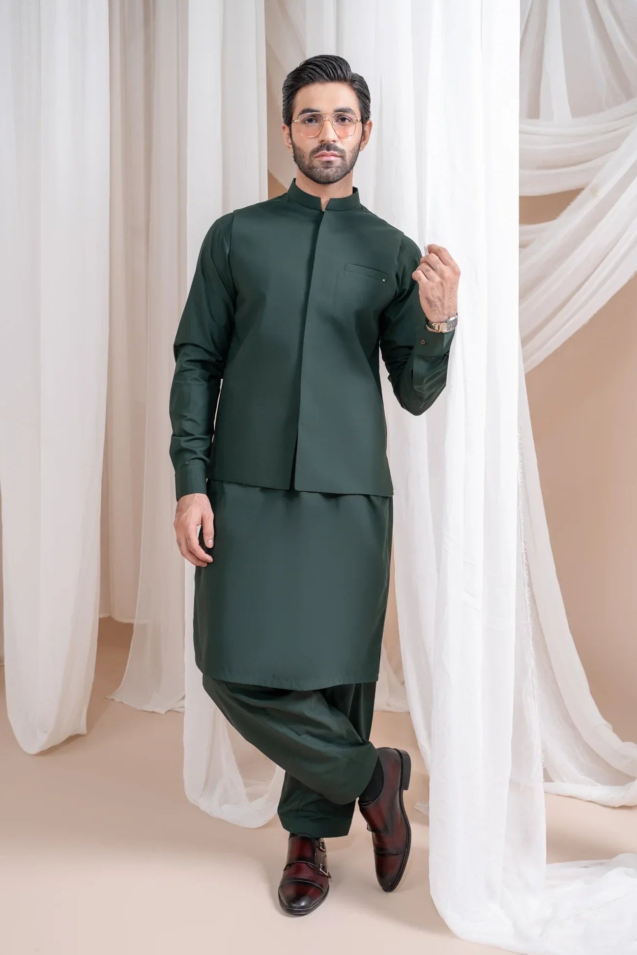 A man wearing a bottle green luxury formal Kameez Shalwar with a patch pocket waistcoat and full sleeves. The outfit is straight cut and suited for all-season wear.