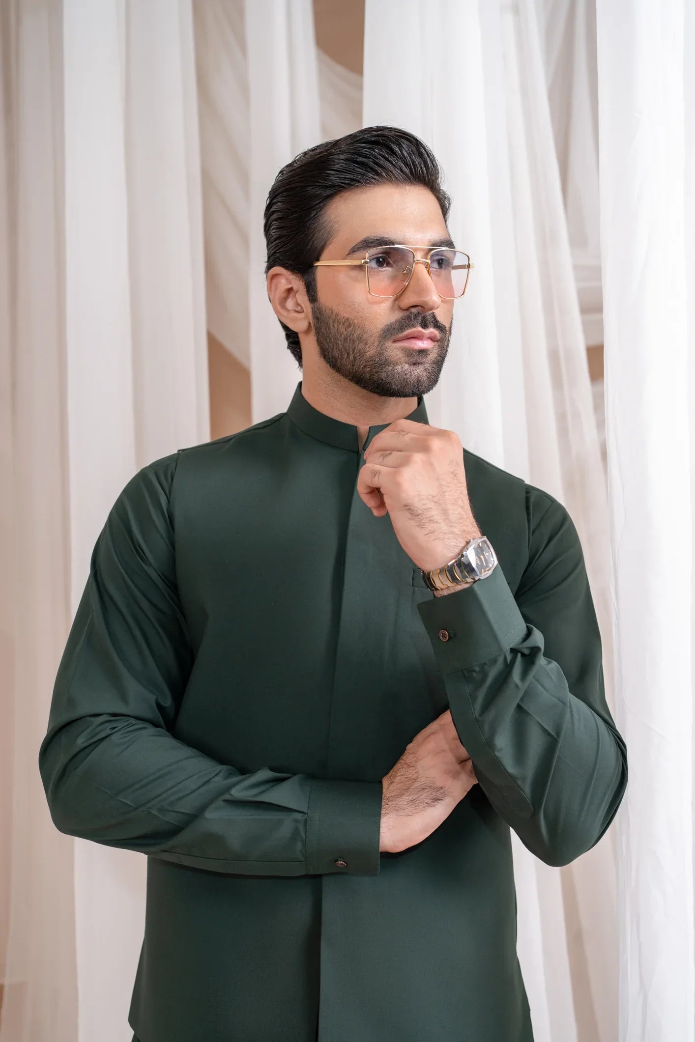 A man wearing a green luxury blended waistcoat with a mandarin collar and straight cut design, standing in a room with white drapes in the background.
