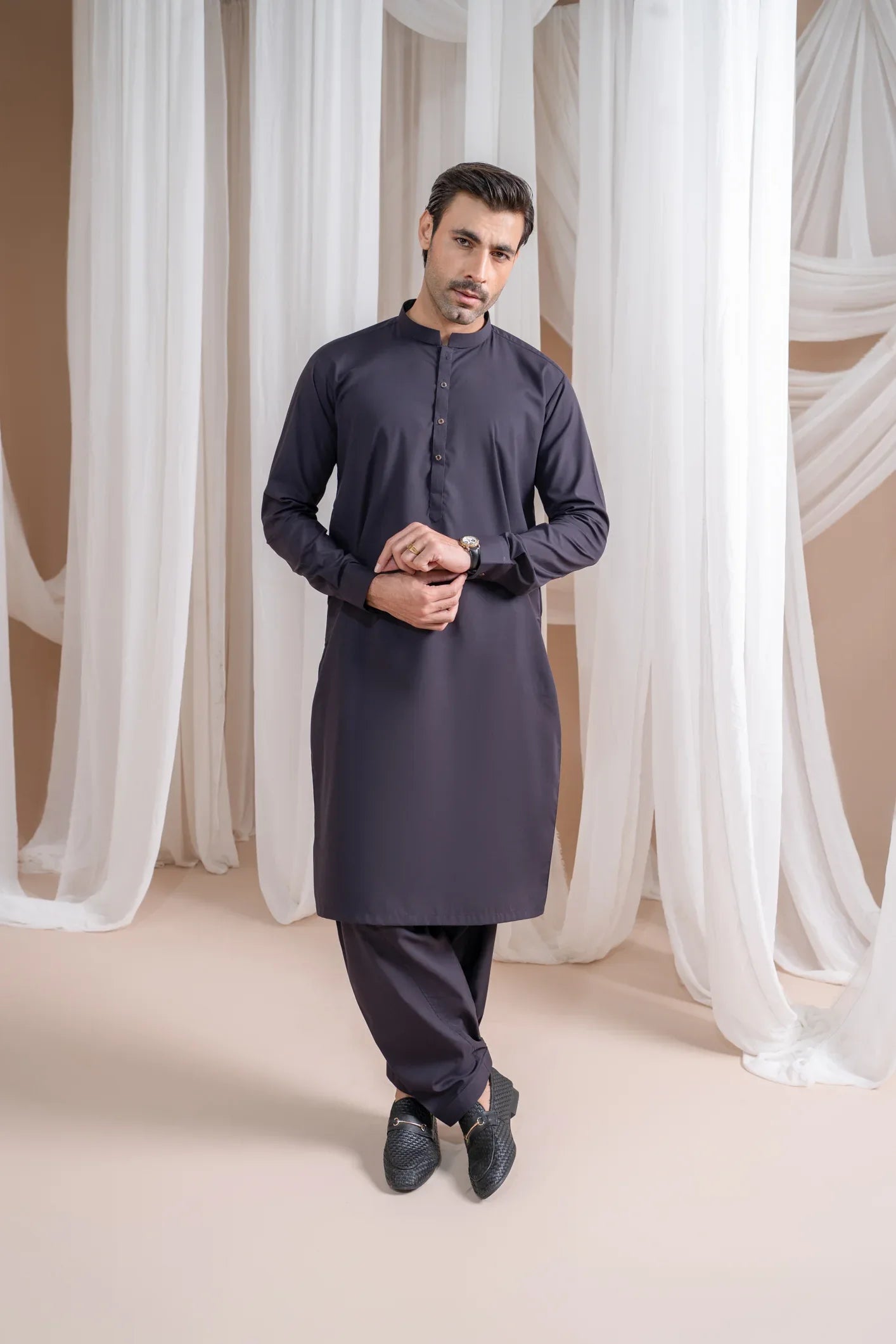 A man wearing a deep burgundy luxury blended kurta shalwar and waistcoat, standing in a room with white drapes in the background.