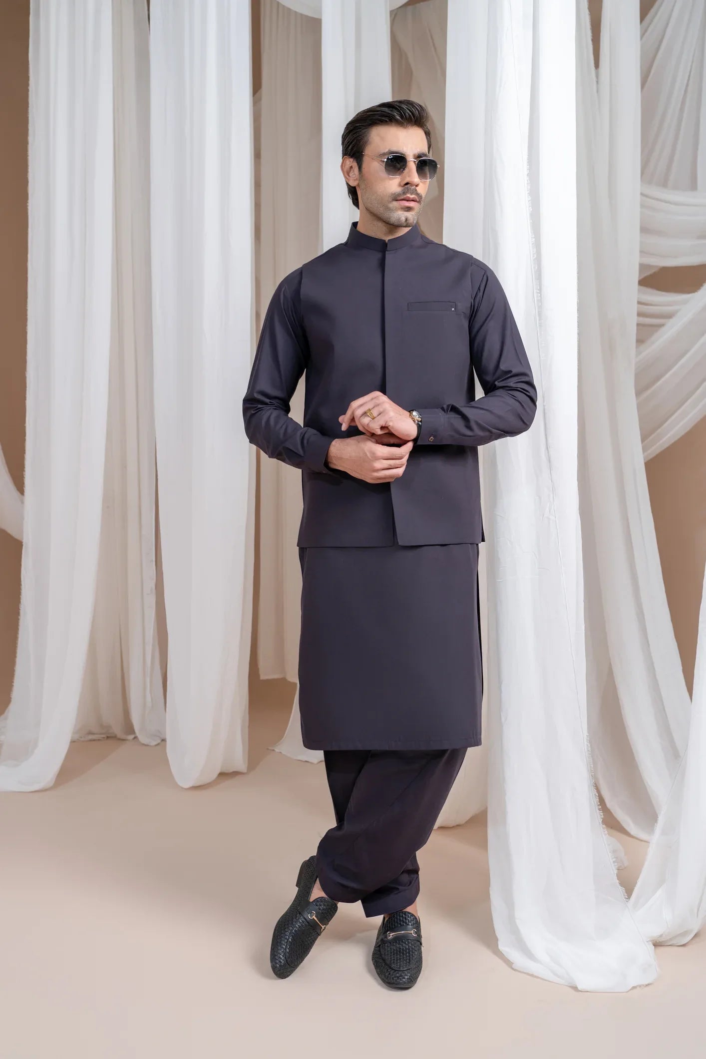 A man wearing a burgundy luxury formal kameez shalwar with a blended waistcoat and full sleeves. He is standing in a room with light curtains behind him.