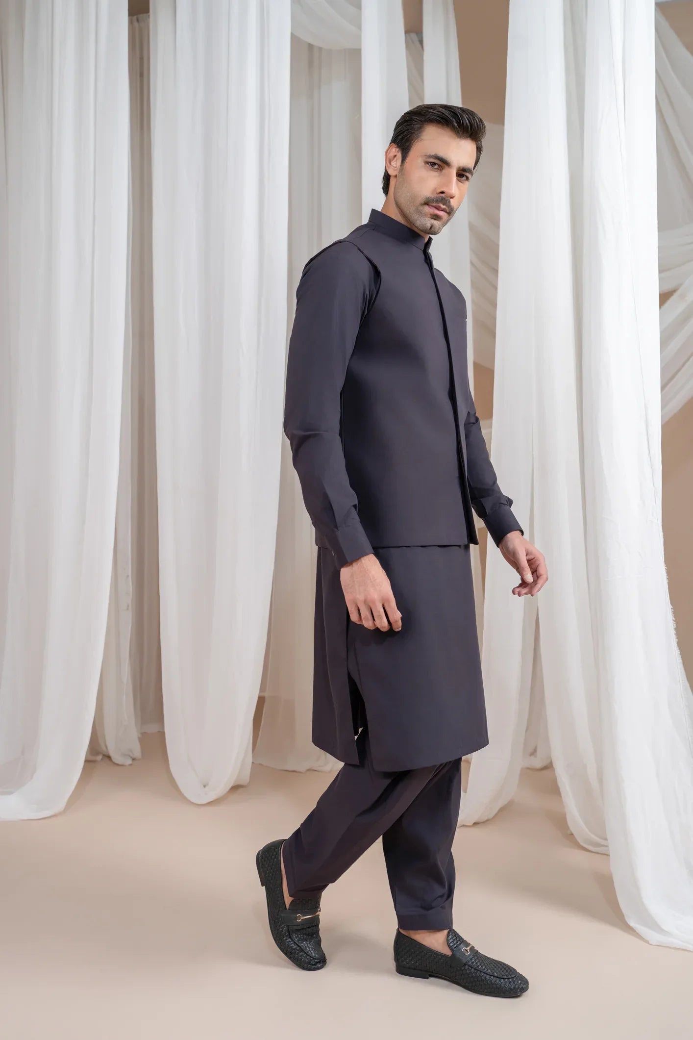 A man wearing a burgundy luxury formal kameez shalwar with a blended waistcoat and full sleeves. He is standing in a room with light curtains behind him.