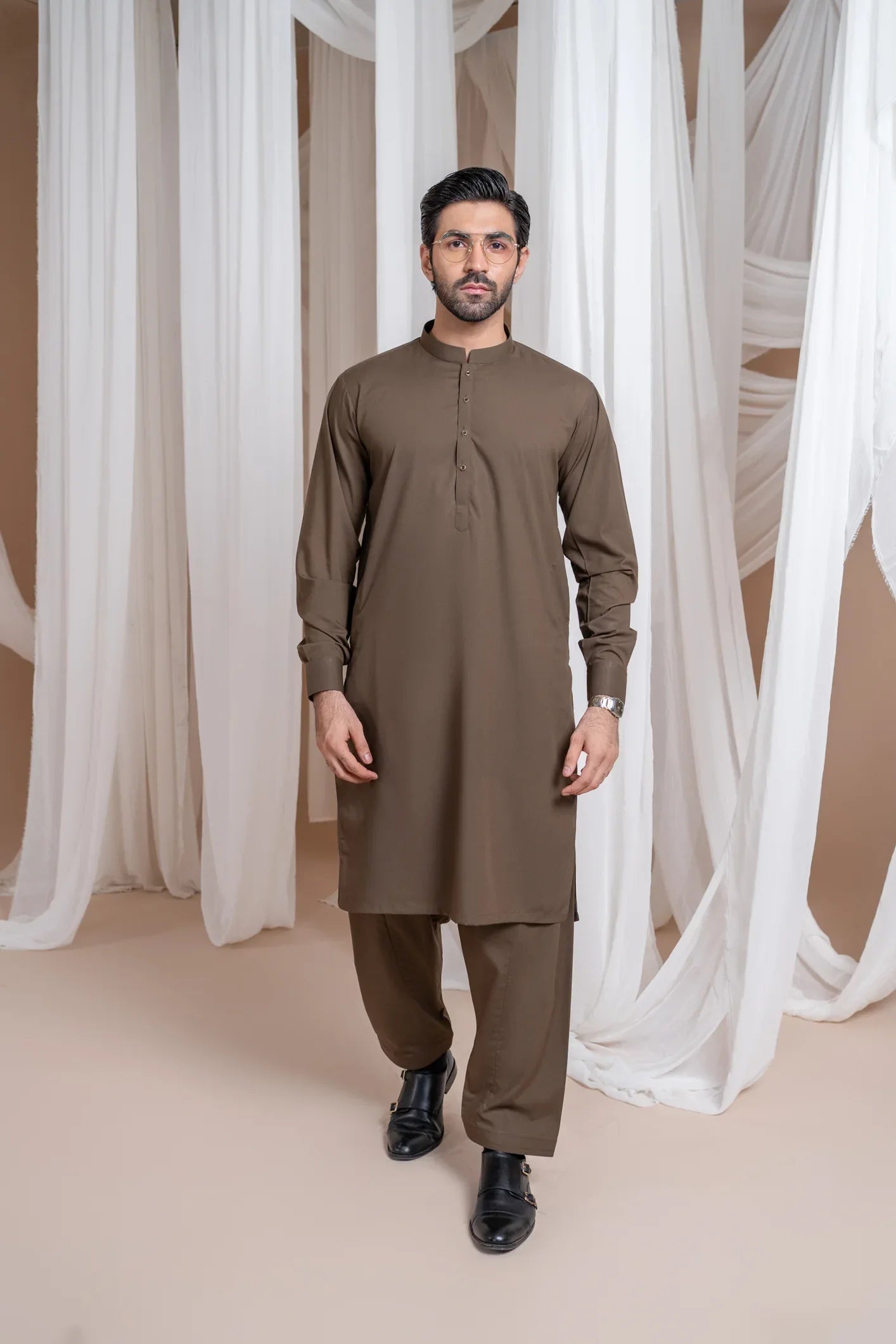 A man wearing a light brown luxury blended kurta shalwar set with a mandarin collar, full sleeves, and a straight kurta and trouser design, standing in a room with white draped backgrounds.