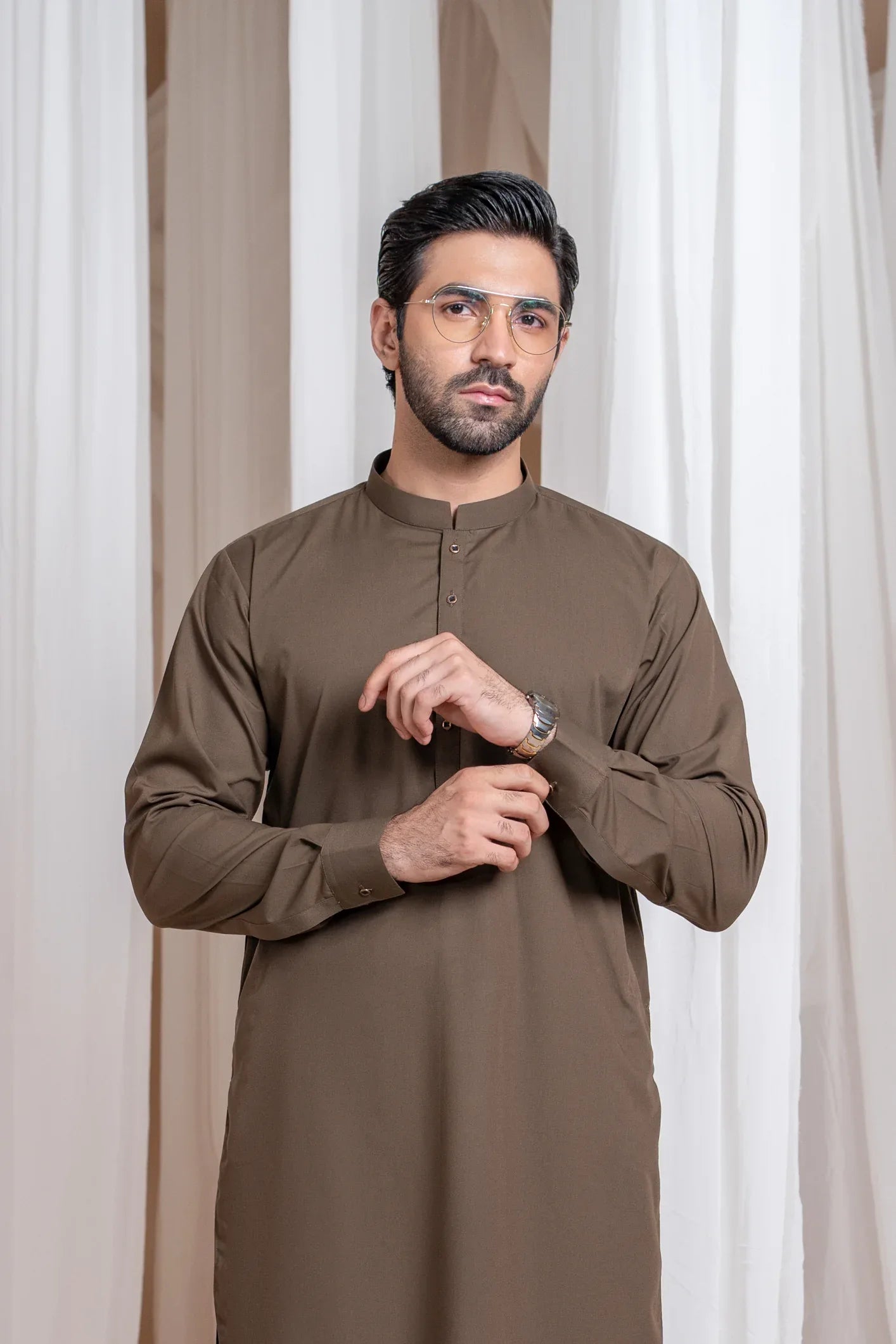 A man wearing a light brown luxury blended kurta shalwar set with a mandarin collar, full sleeves, and a straight kurta and trouser design, standing in a room with white draped backgrounds.