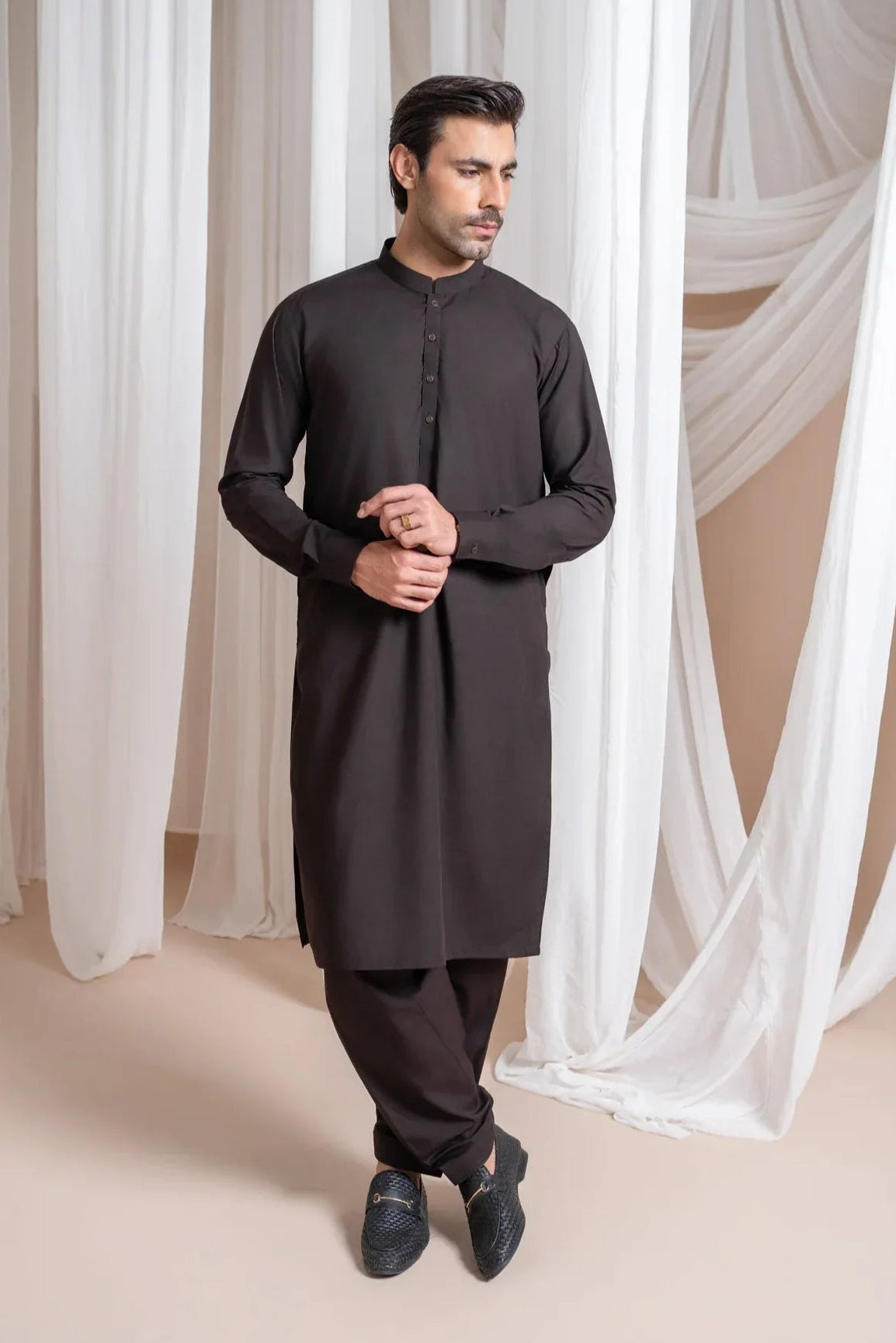 A man wearing a dark brown kameez shalwar with a mandarin collar and full sleeves, standing in a room with white drapes in the background.