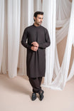 A man wearing a dark brown kameez shalwar with a mandarin collar and full sleeves, standing in a room with white drapes in the background.