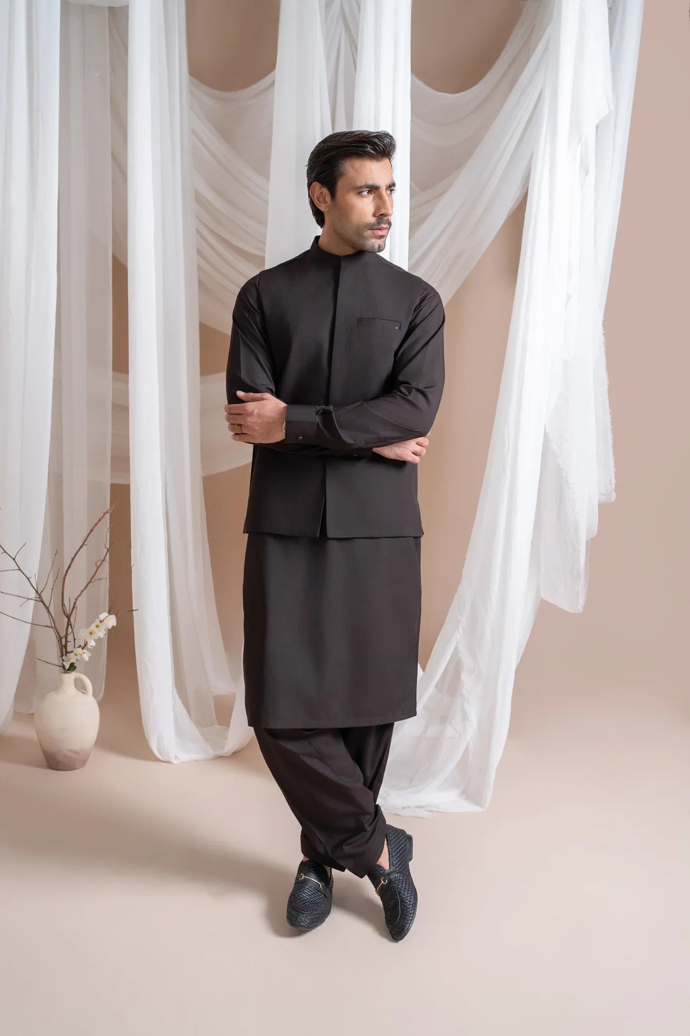 A man wearing a dark brown straight cut kameez shalwar with a blended waistcoat standing indoors with curtains in the background.