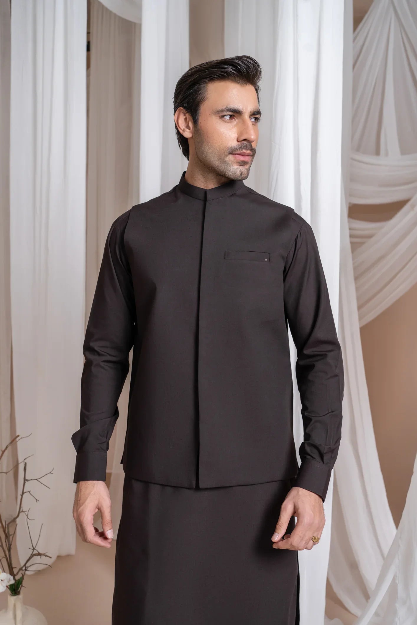 A man wearing a dark brown straight cut kameez shalwar with a blended waistcoat standing indoors with curtains in the background.