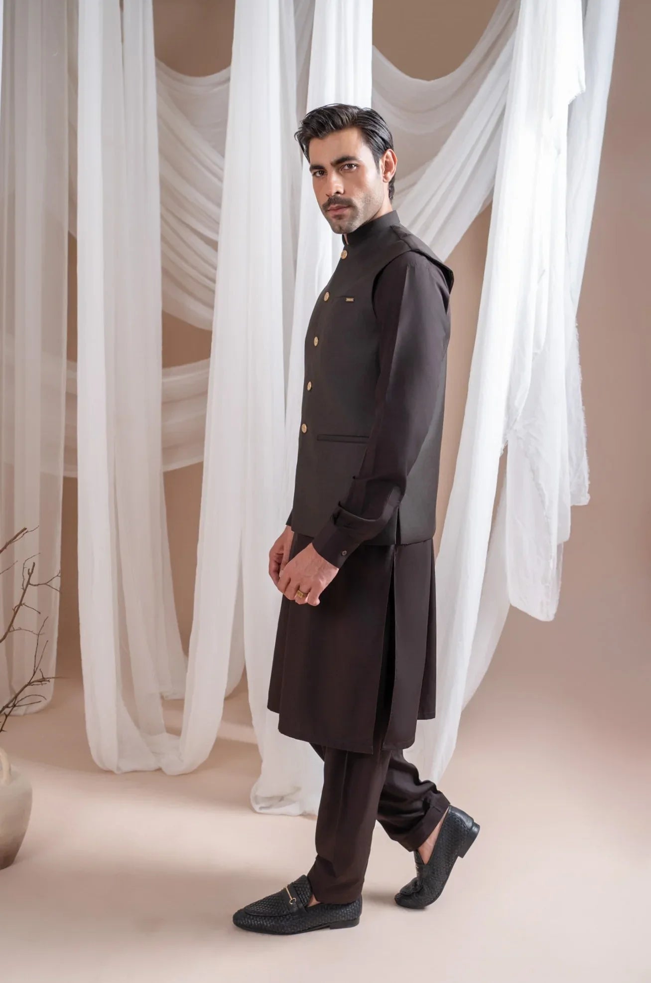 Man wearing a dark brown shalwar kameez and waistcoat with white curtains in the background