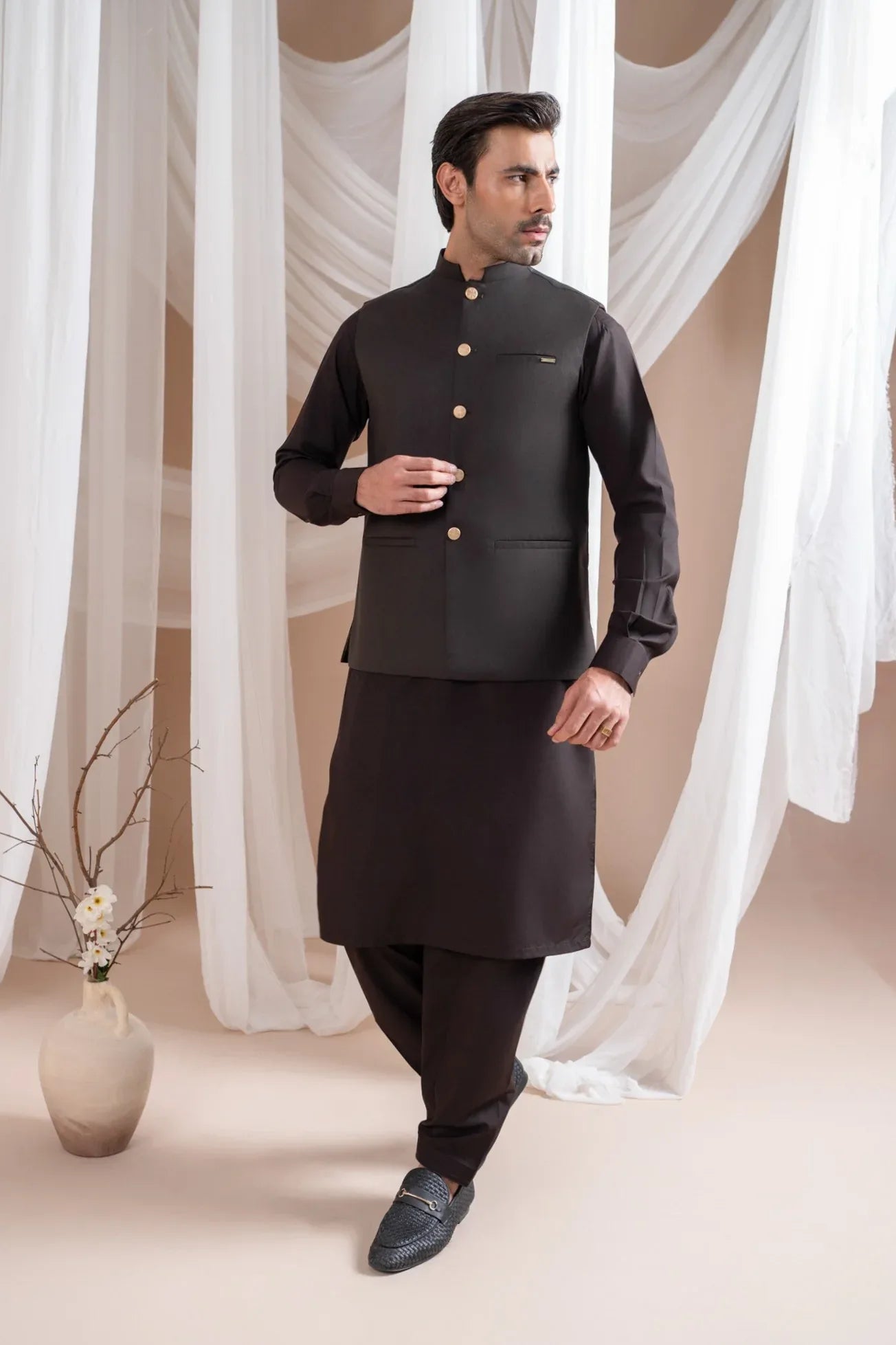 Man wearing a dark brown shalwar kameez and waistcoat with white curtains in the background