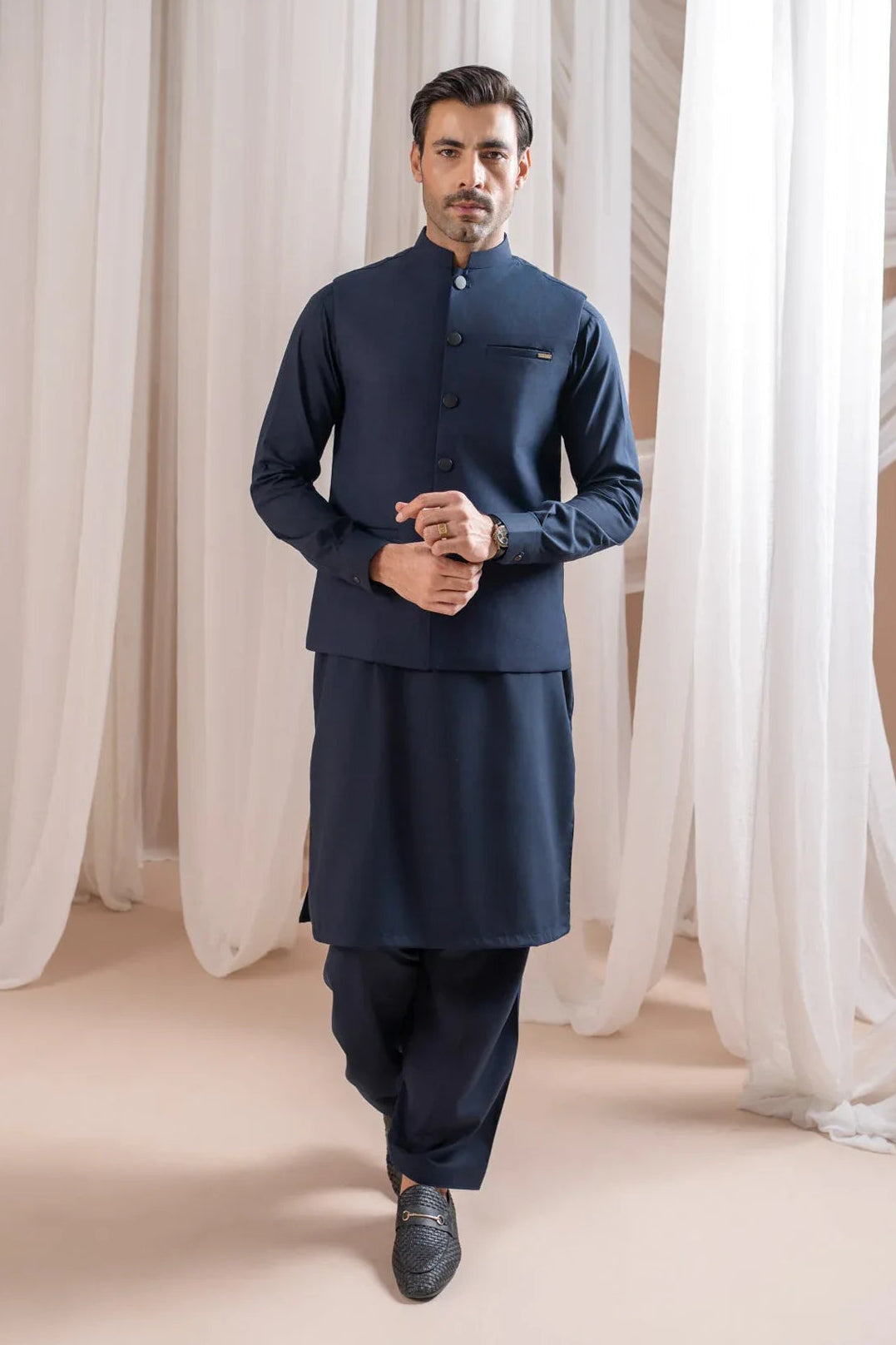 Man wearing a navy blue shalwar kameez with navy blue suiting waistcoat against a white curtain background