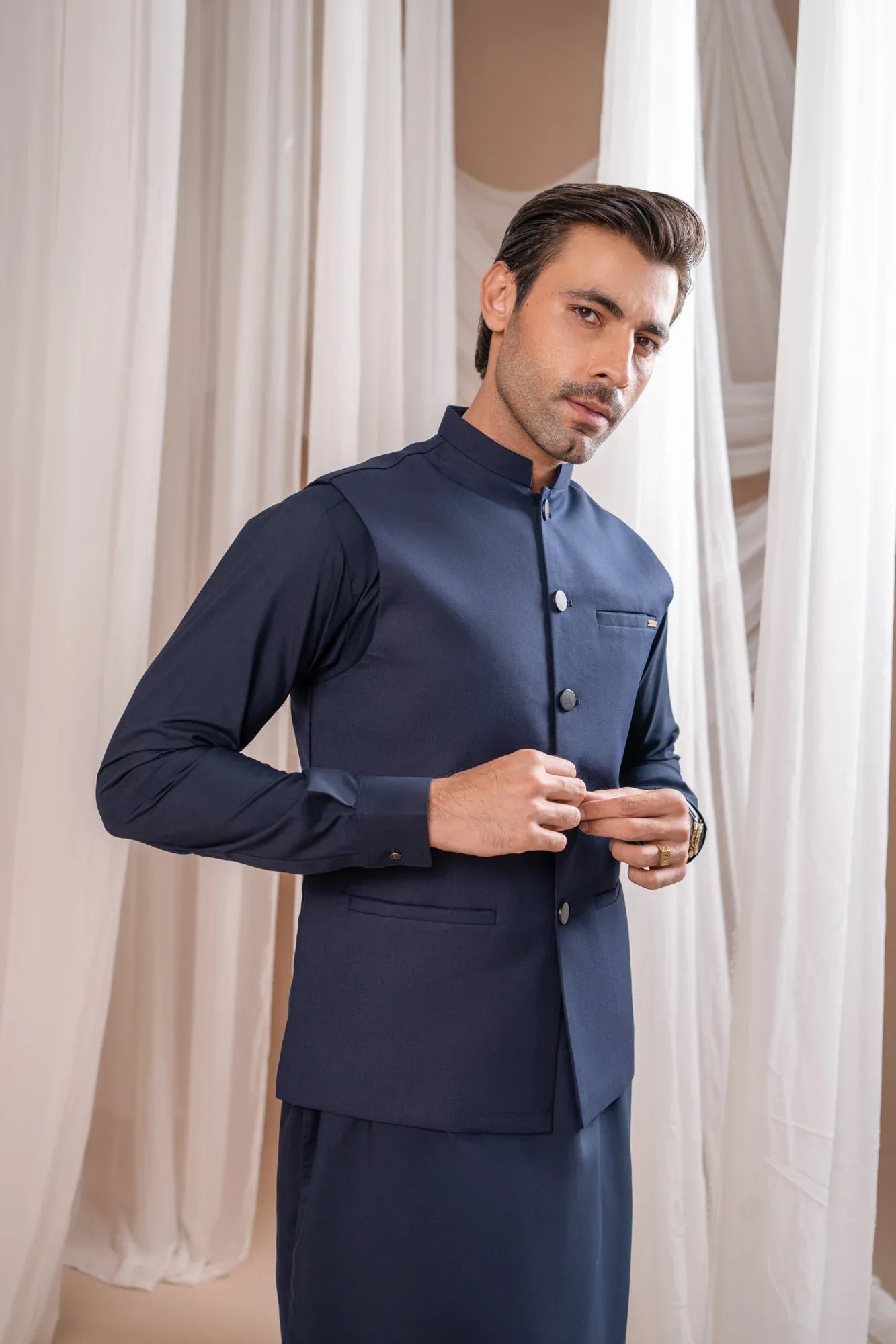 Man wearing a navy blue shalwar kameez with navy blue suiting waistcoat against a white curtain background