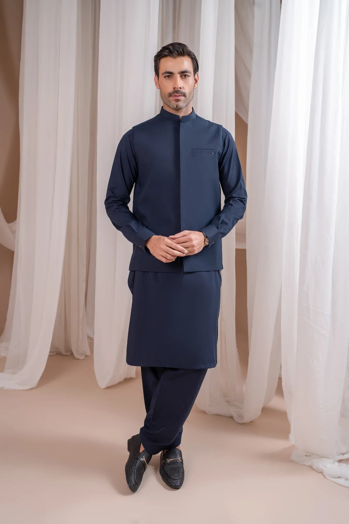 A man wearing a navy blue formal kameez shalwar with a blended waistcoat and full sleeves. He is standing against a neutral backdrop.
