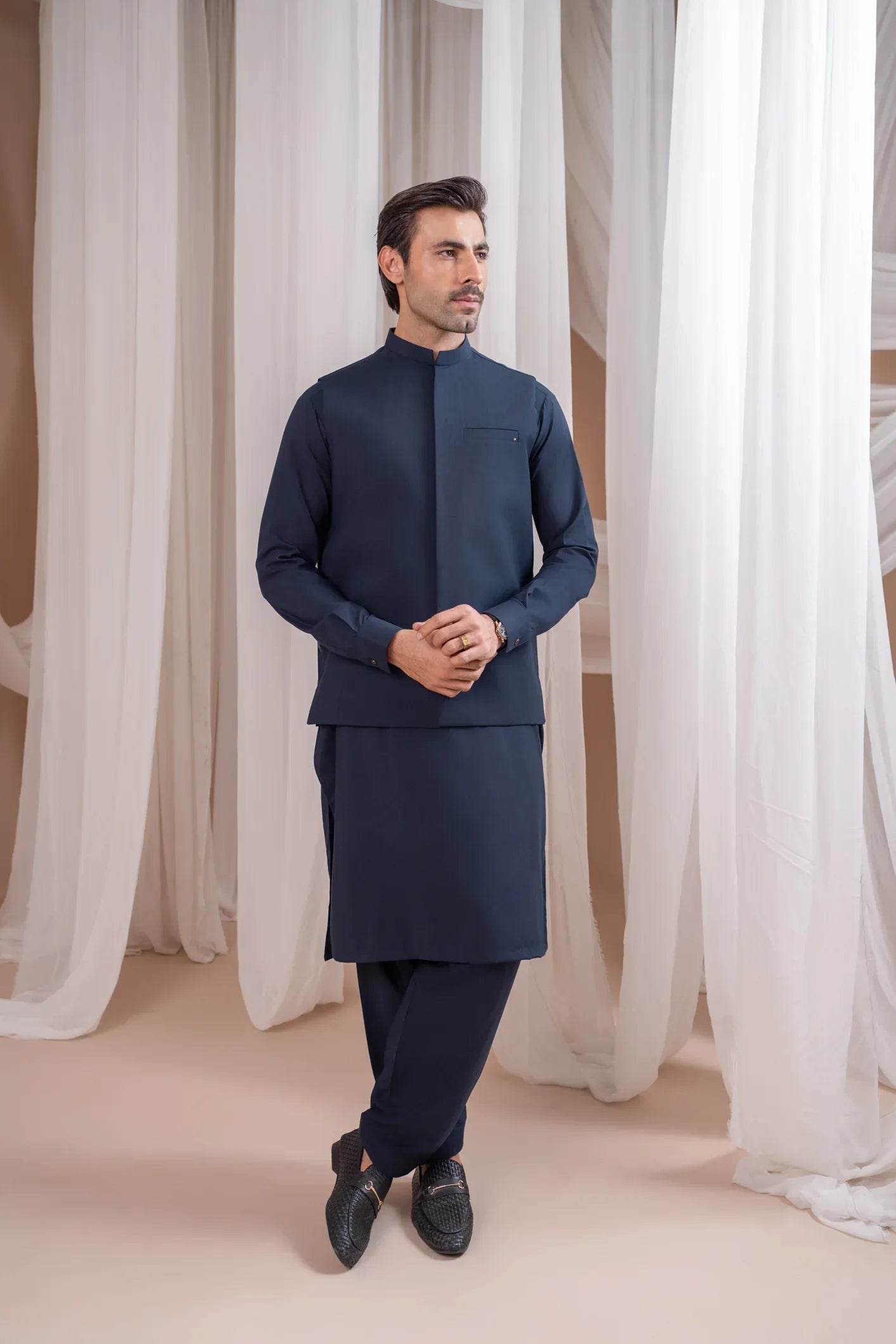 A man wearing a navy blue formal kameez shalwar with a blended waistcoat and full sleeves. He is standing against a neutral backdrop.