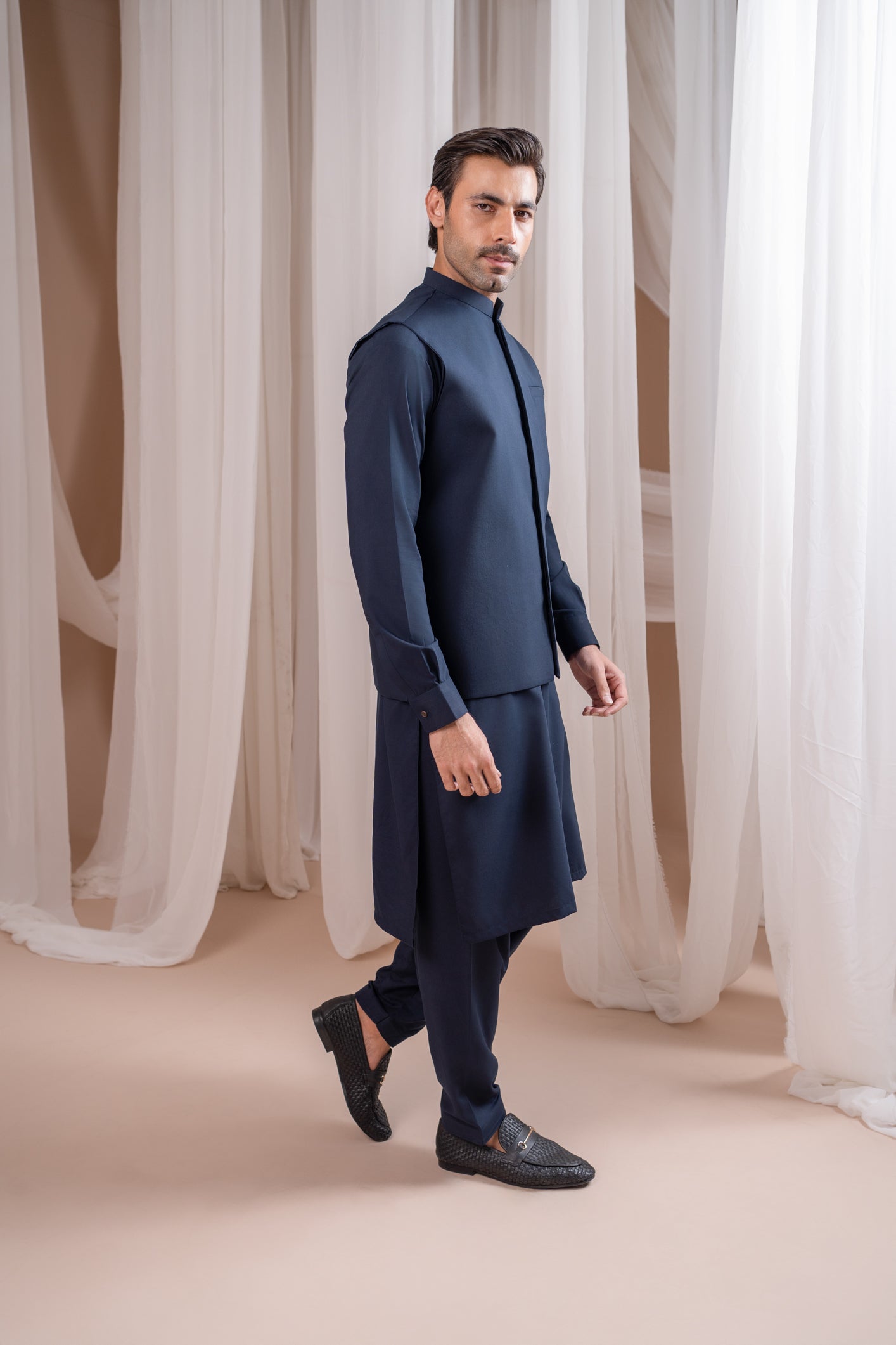 A man wearing a navy blue formal kameez shalwar with a blended waistcoat and full sleeves. He is standing against a neutral backdrop.