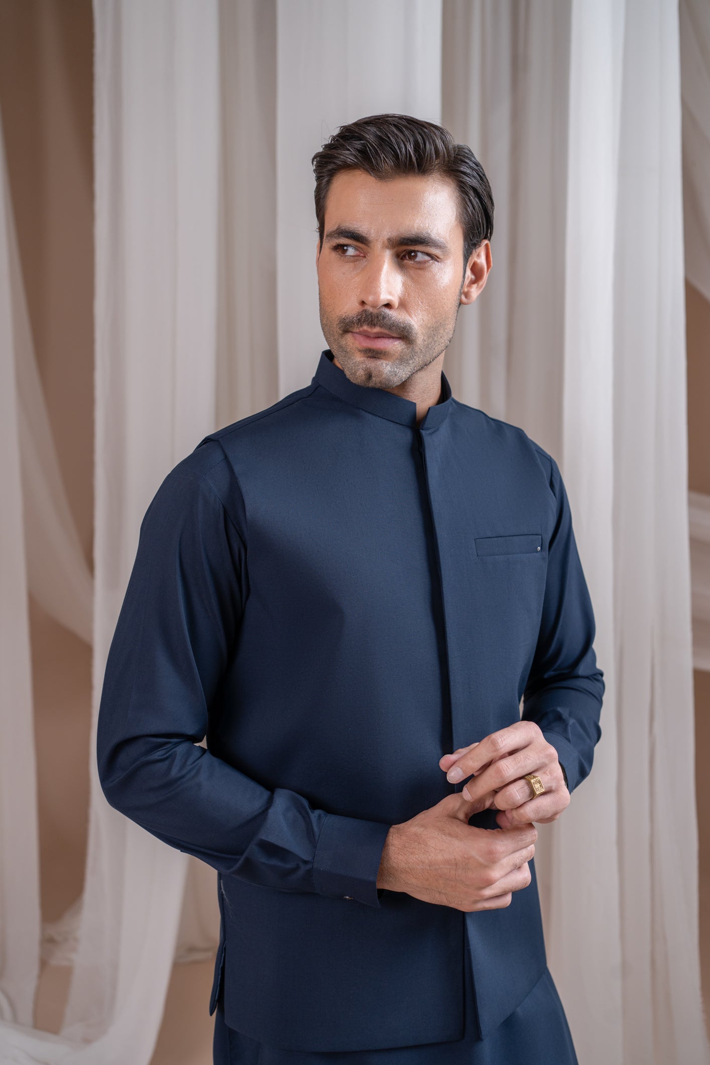 A man wearing a navy blue formal kameez shalwar with a blended waistcoat and full sleeves. He is standing against a neutral backdrop.