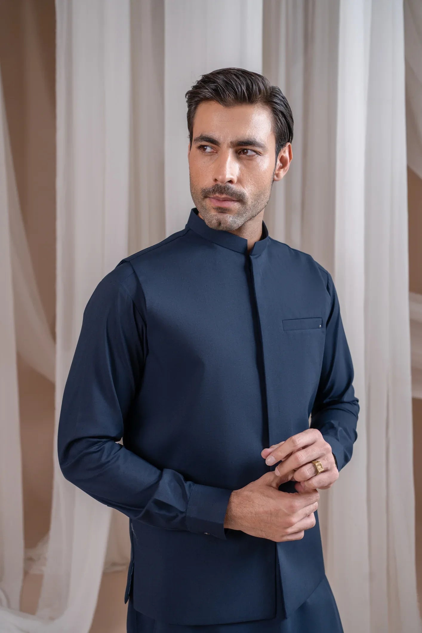 A man wearing a navy blue formal kameez shalwar with a blended waistcoat and full sleeves. He is standing against a neutral backdrop.