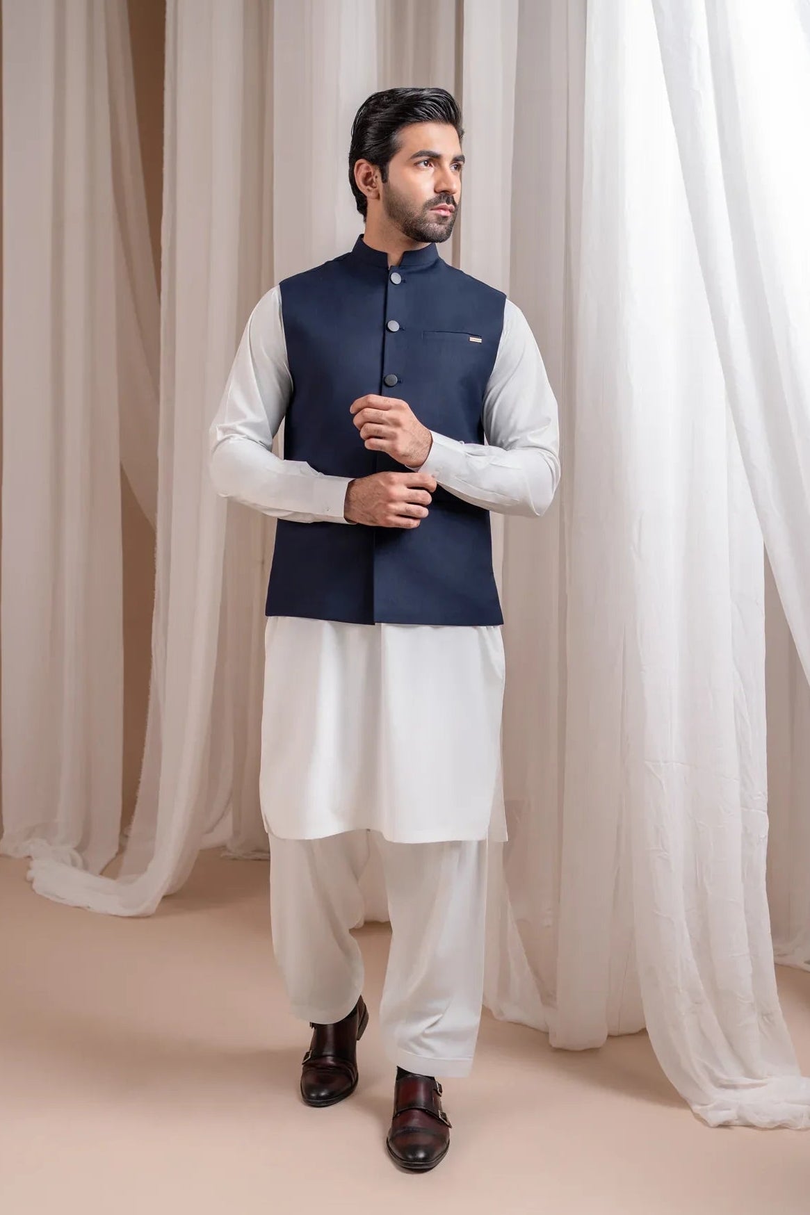 Man wearing an off white shalwar kameez with a navy blye waistcoat against white curtains