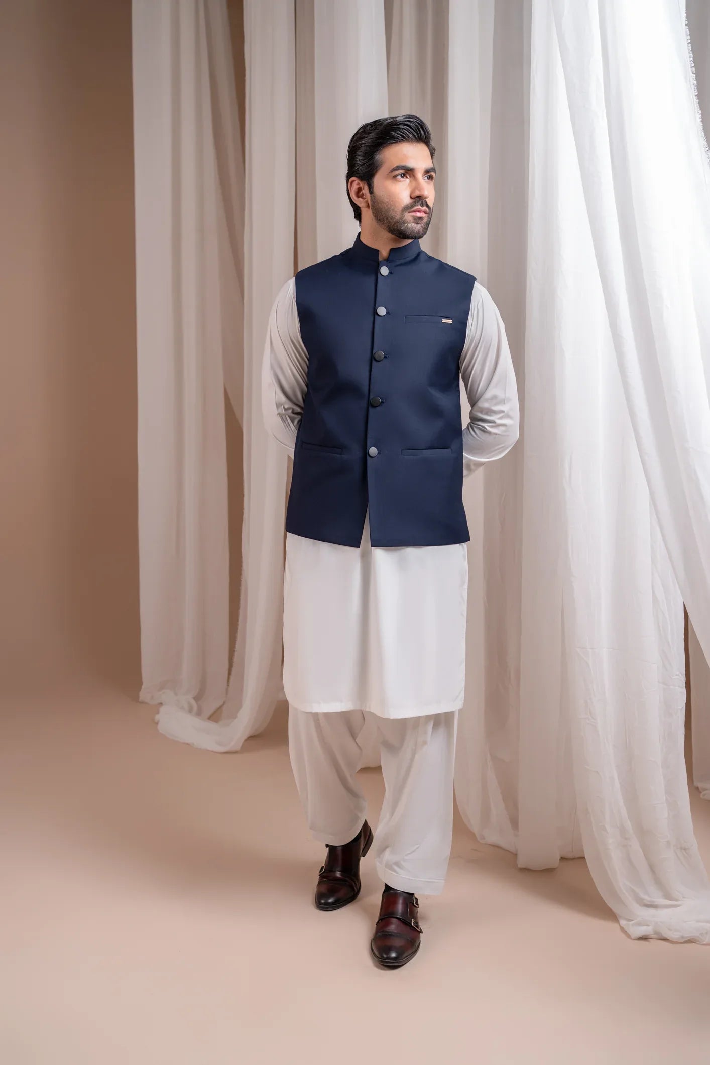 Man wearing an off white shalwar kameez with a navy blye waistcoat against white curtains