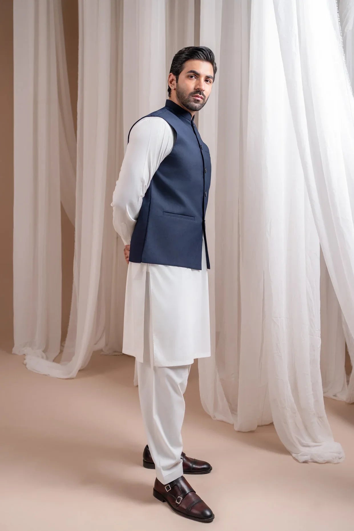 Man wearing an off white shalwar kameez with a navy blye waistcoat against white curtains