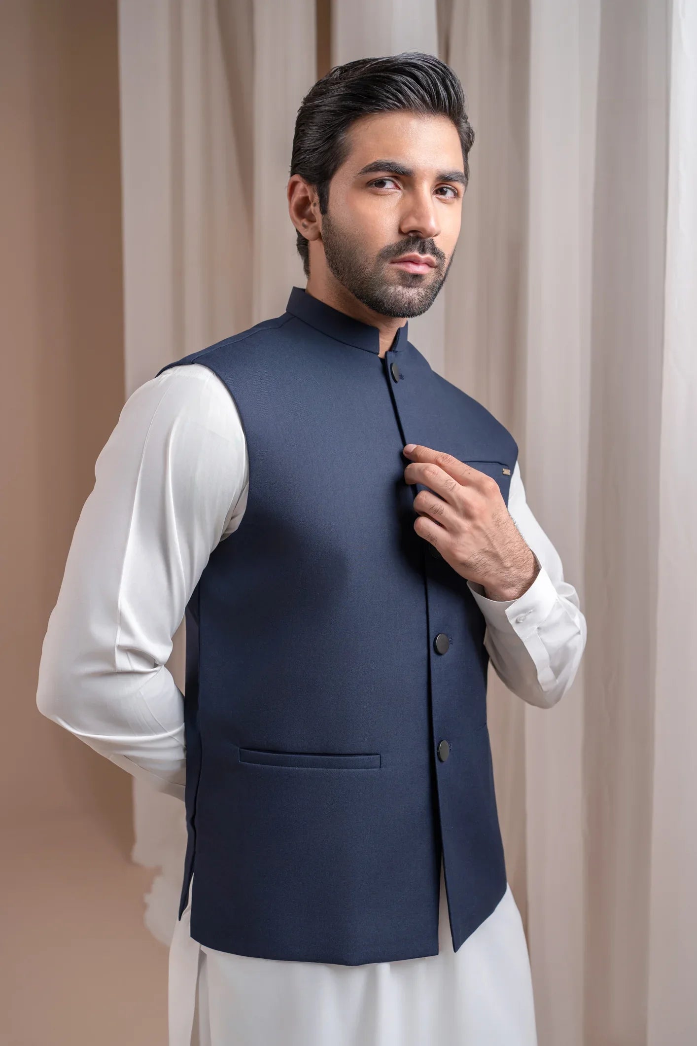 Man wearing an off white shalwar kameez with a navy blye waistcoat against white curtains
