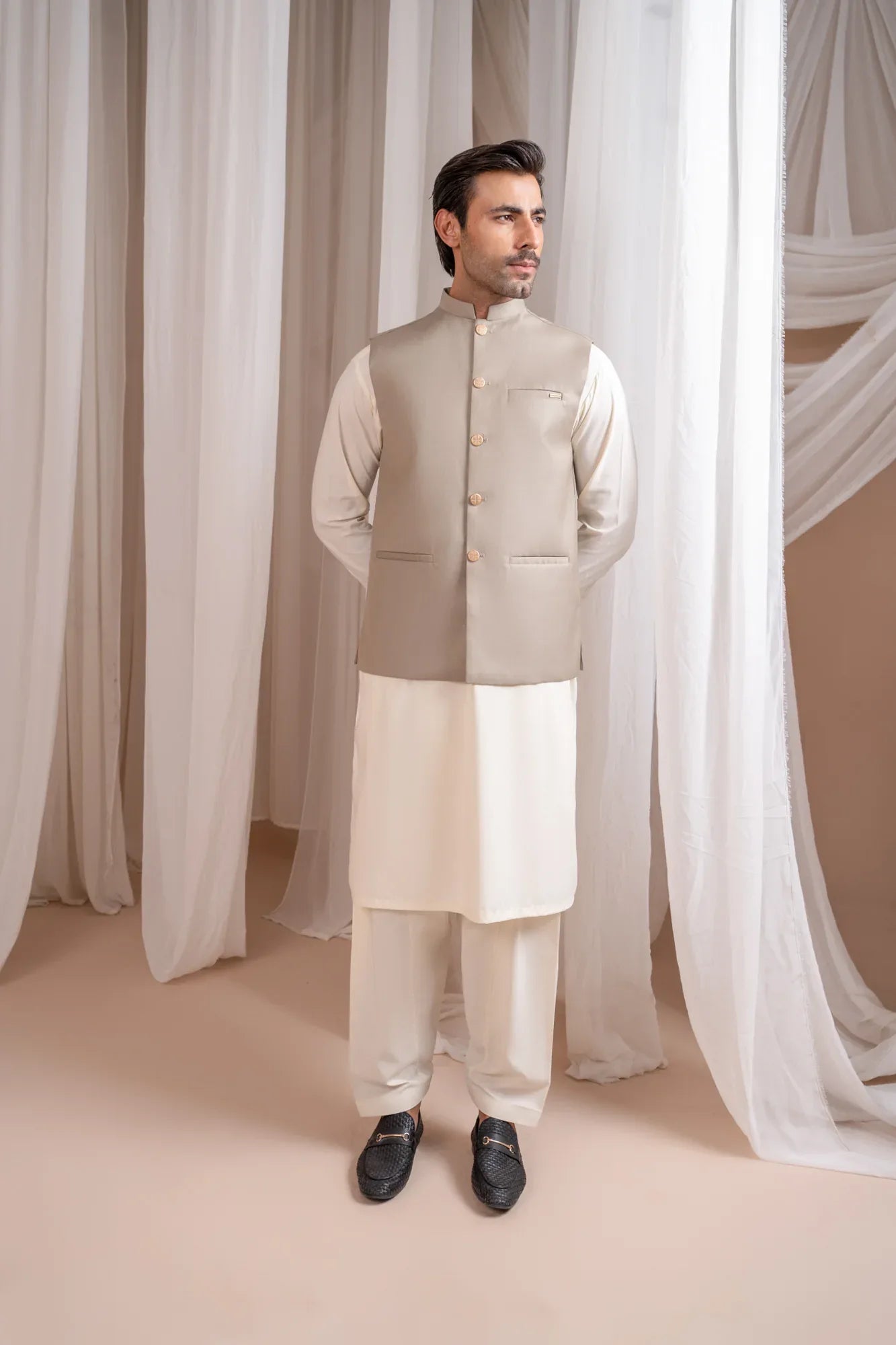 Man wearing a beige suiting waistcoat and white shalwar kameez against a white curtain background