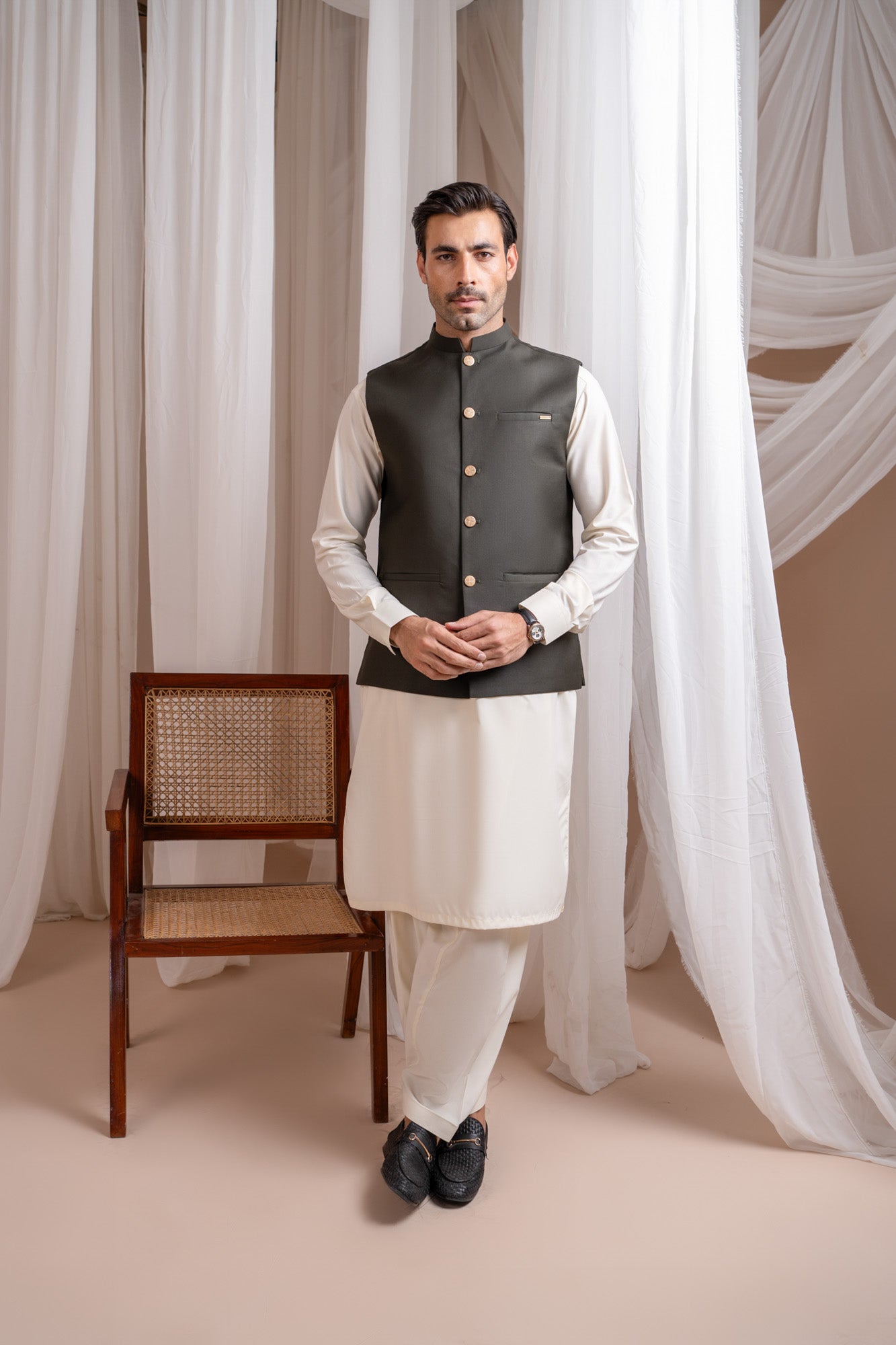 Man wearing a army green suiting waistcoat over bosky white kameez shalwar with a neutral background