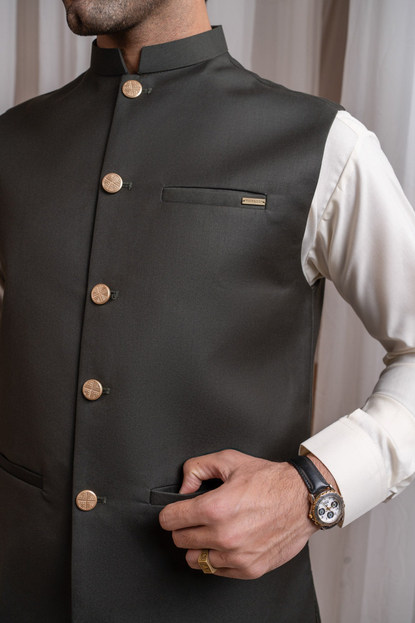 Man wearing a army green suiting waistcoat over bosky white kameez shalwar with a neutral background