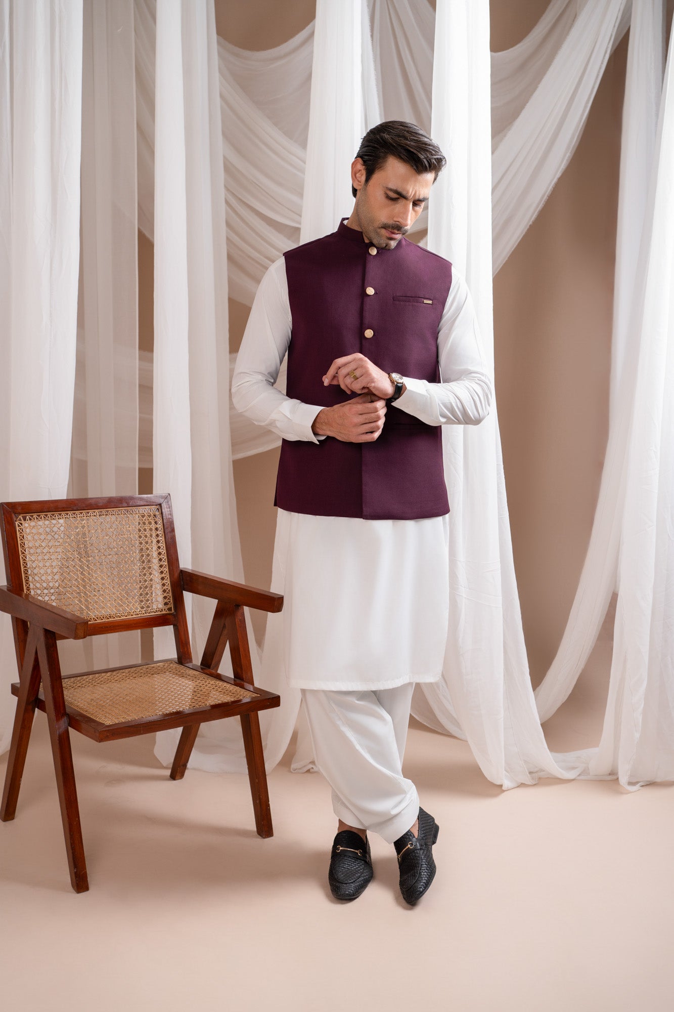 Man wearing a burgundy suitinf waistcoat over an off white kameez shalwar, standing in a room with curtains and a chair.