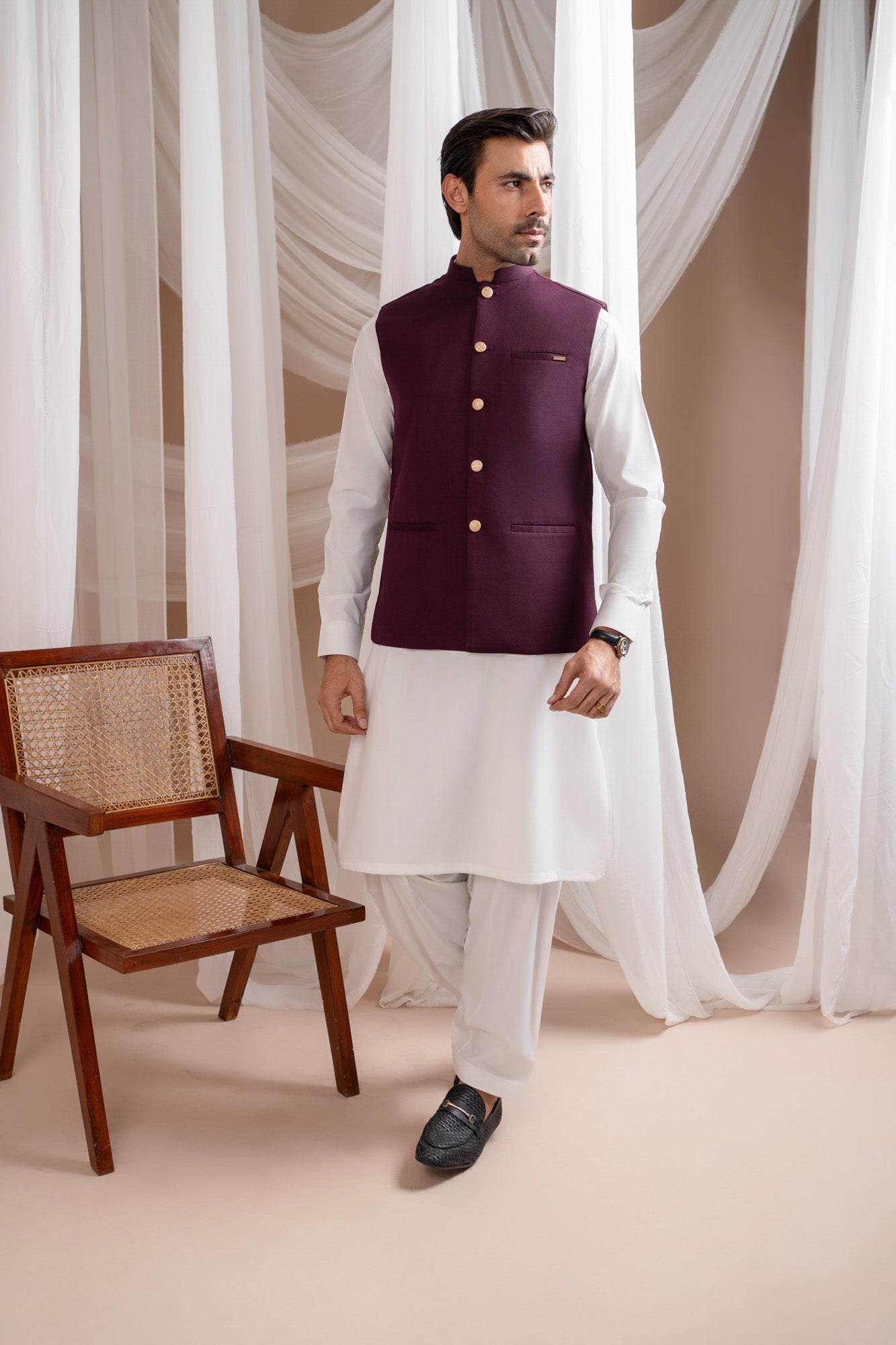 Man wearing a burgundy suitinf waistcoat over an off white kameez shalwar, standing in a room with curtains and a chair.