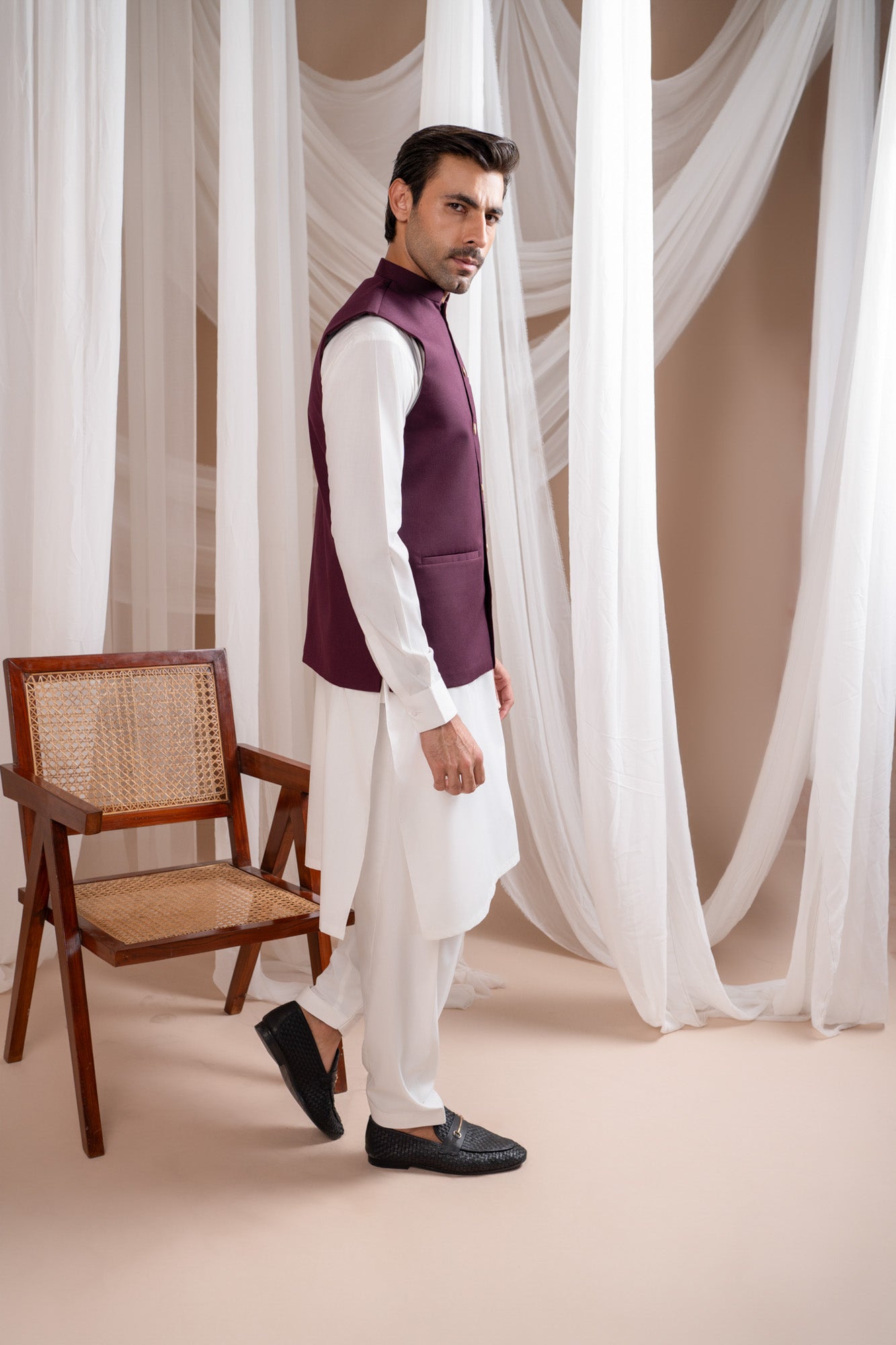 Man wearing a burgundy suitinf waistcoat over an off white kameez shalwar, standing in a room with curtains and a chair.
