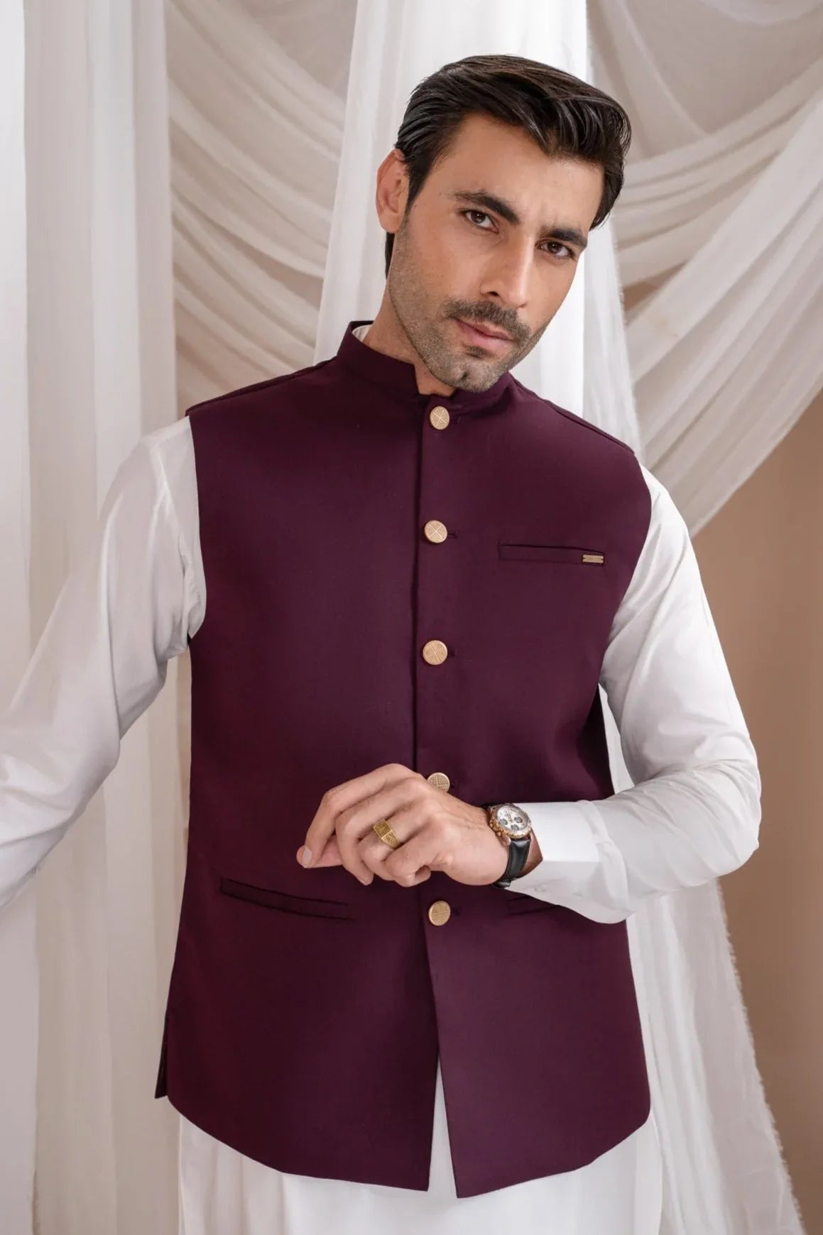 A man wearing a straight cut, buttoned, burgundy suiting waistcoat over a white shirt.