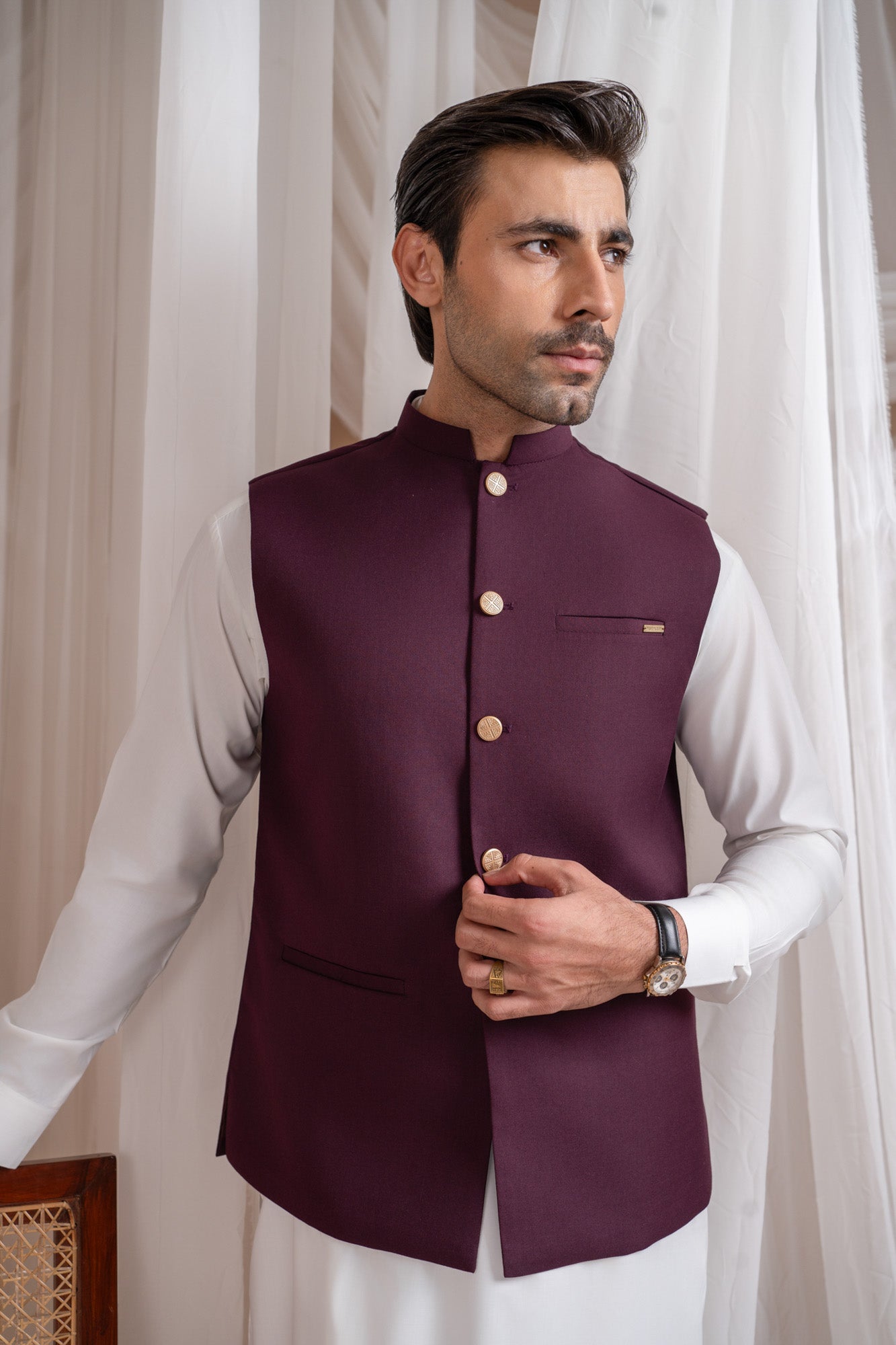 Man wearing a burgundy suitinf waistcoat over an off white kameez shalwar, standing in a room with curtains and a chair.