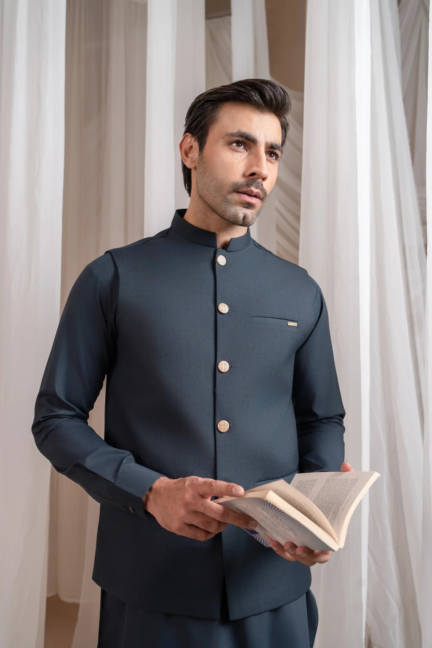 A man standing in a traditional attire, consisting of a charcoal grey kurta shalwar and a matching suiting waistcoat, against a backdrop with white drapes.