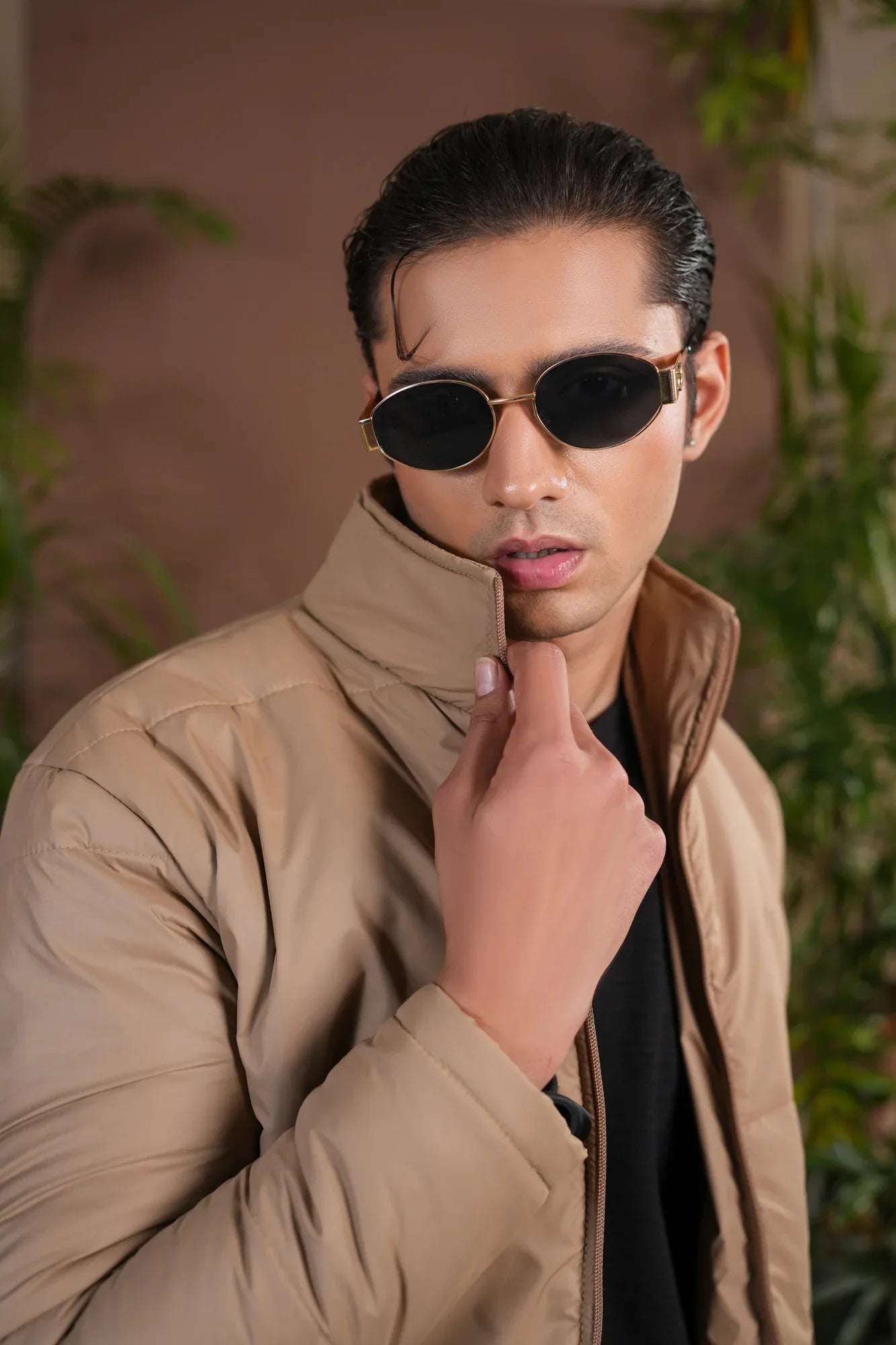 Man wearing sunglasses and a beige jacket with plants in the background