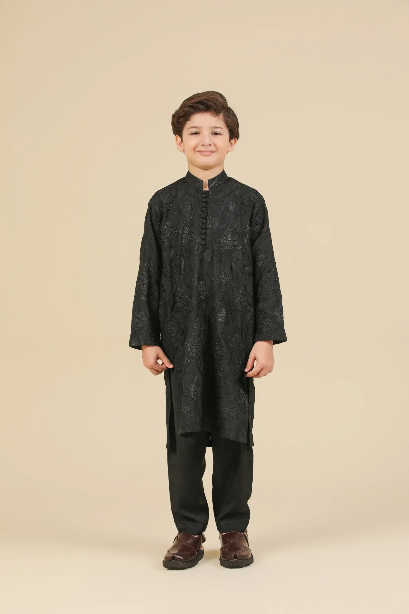 Young boy wearing a black raw silk embroidered kurta trouser outfit on a beige background