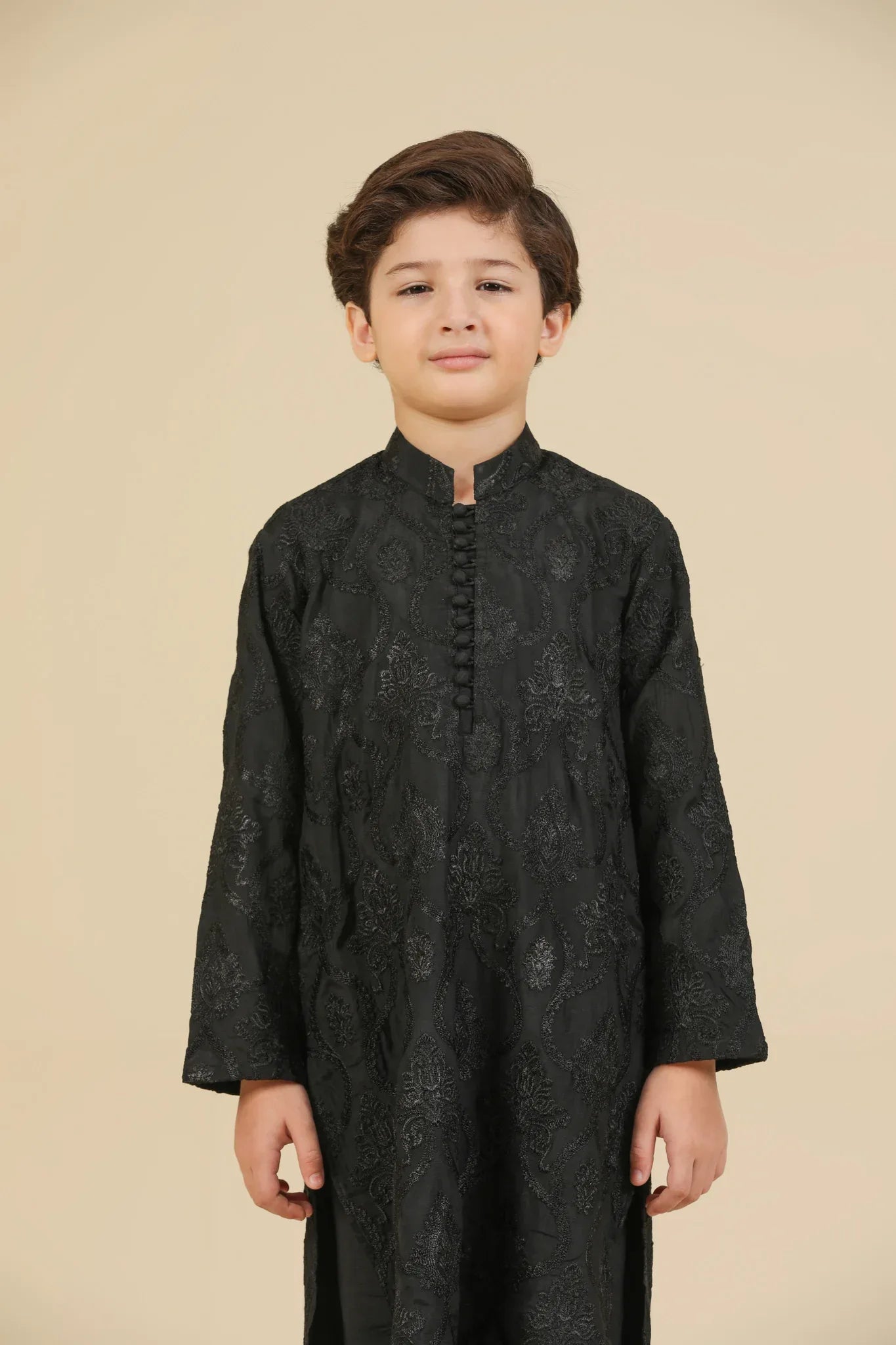 Young boy wearing a black raw silk embroidered kurta trouser outfit on a beige background