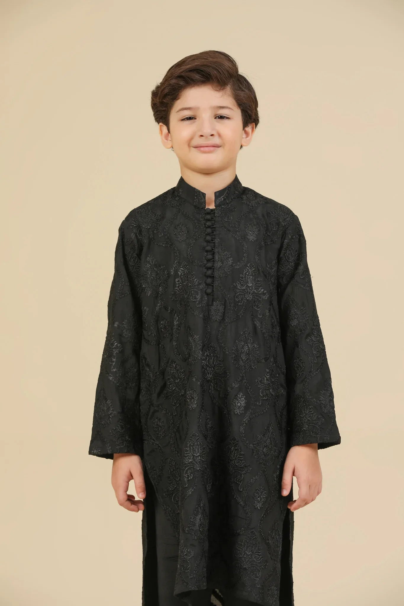 Young boy wearing a black raw silk embroidered kurta trouser outfit on a beige background