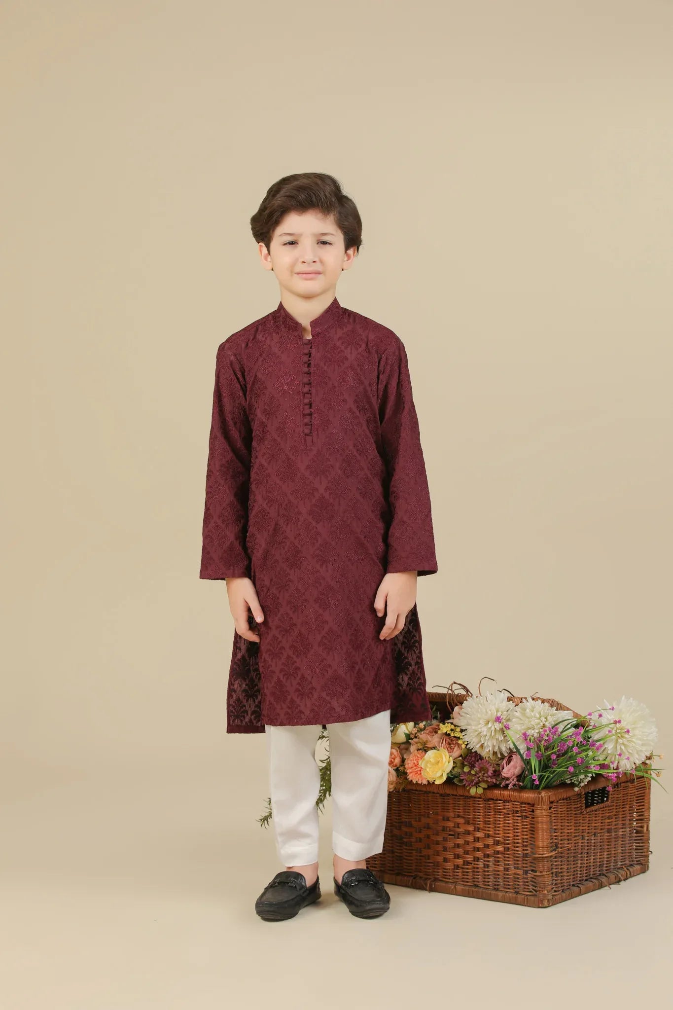 Child wearing a maroon raw silk embroidered kurta with white pants standing next to a basket of flowers on a beige background