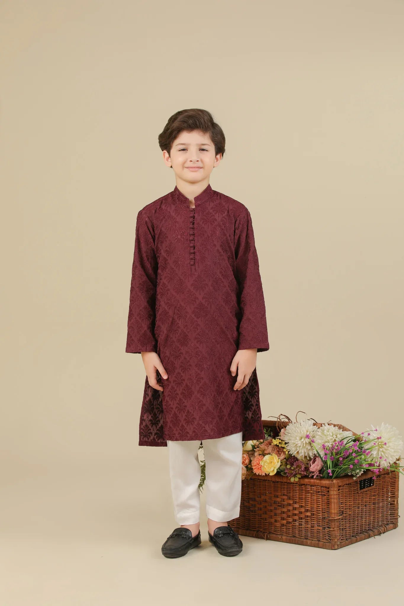 Child wearing a maroon raw silk embroidered kurta with white pants standing next to a basket of flowers on a beige background
