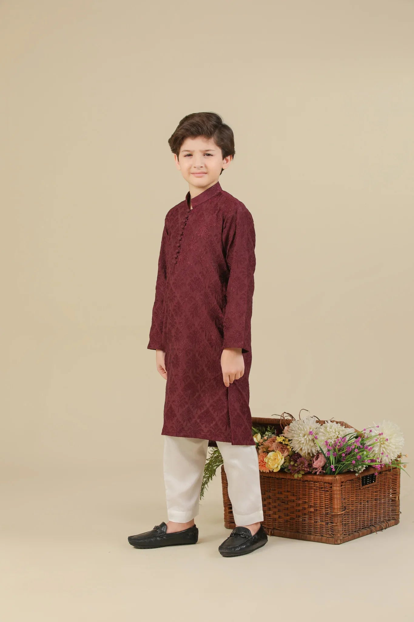Child wearing a maroon raw silk embroidered kurta with white pants standing next to a basket of flowers on a beige background