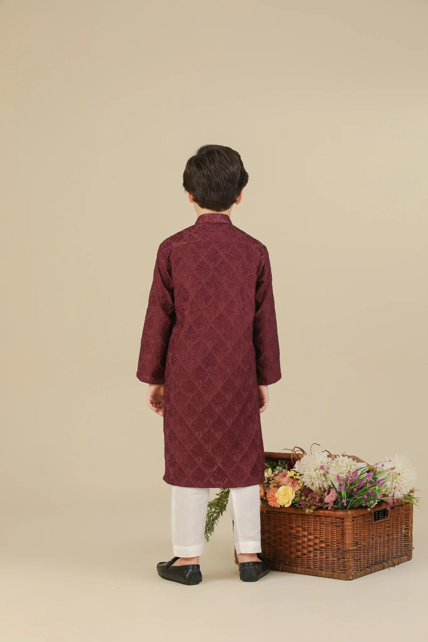 Child wearing a maroon raw silk embroidered kurta with white pants standing next to a basket of flowers on a beige background
