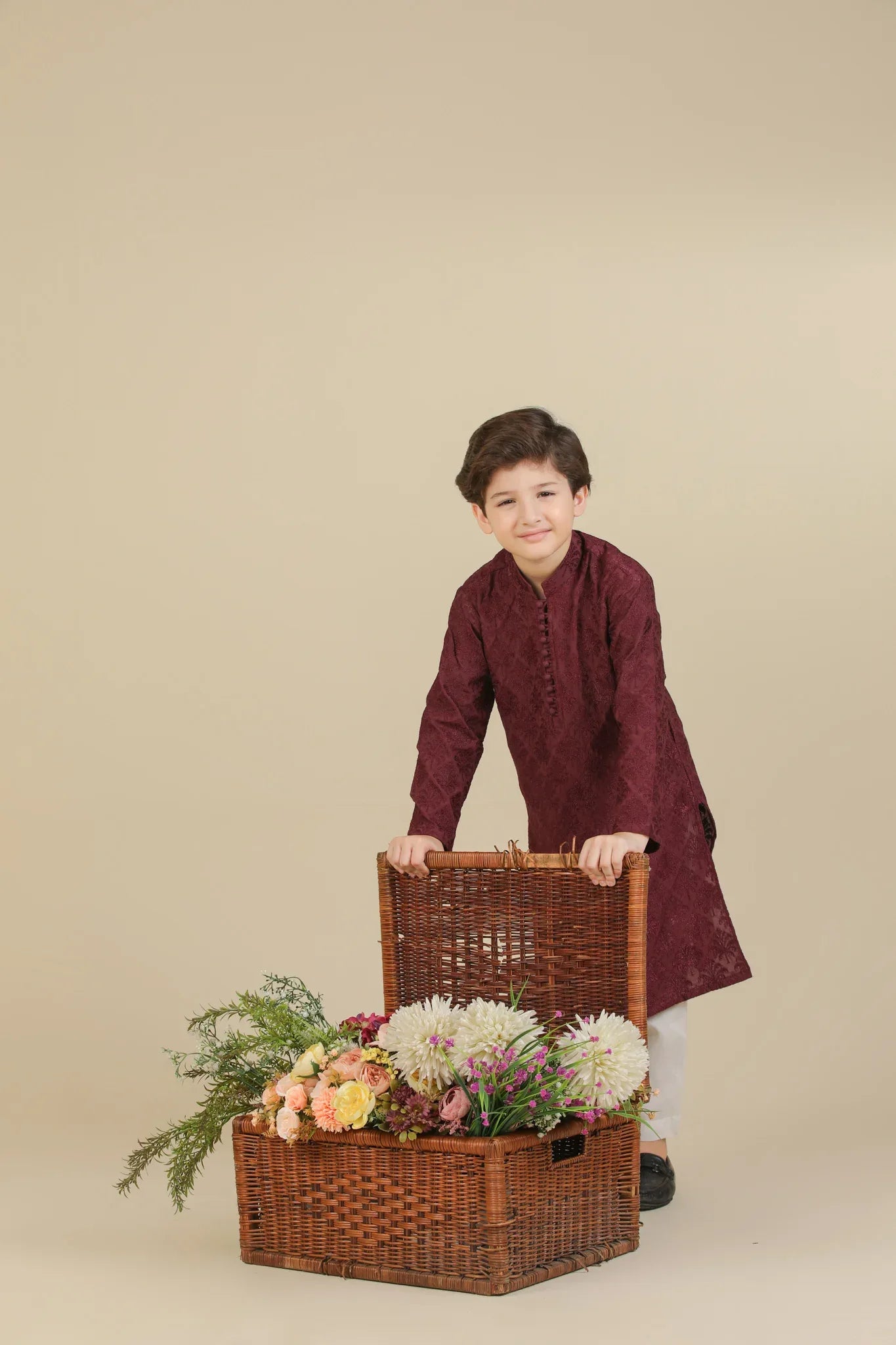 Child wearing a maroon raw silk embroidered kurta with white pants standing next to a basket of flowers on a beige background